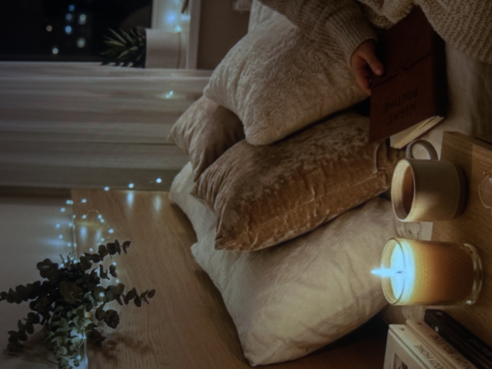 A person is in bed, holding a book. The bed has several pillows, some of which are fluffy and white, and one that is a textured brown. A lit candle sits on a wooden surface next to a mug of tea. In the background, sheer white curtains are visible, and beyond them, the lights of a city at night can be seen. There are also string lights draped around some eucalyptus branches above the bed. This scene is likely in a bedroom in Knezha, Bulgaria, given the context provided.