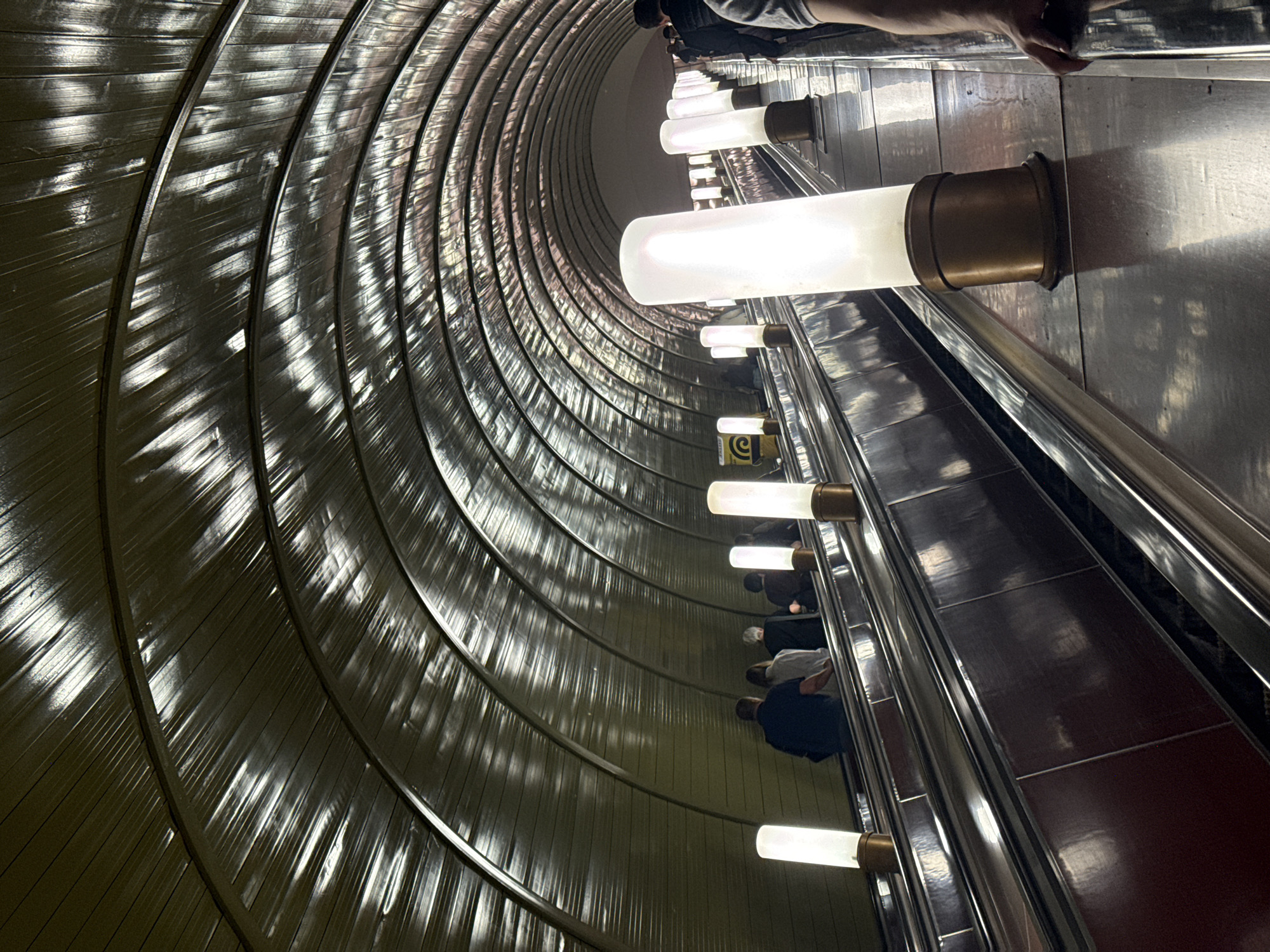 This vertical shot captures the descent down a long, curved escalator tunnel within a Moscow metro station. The perspective looks steeply downwards, emphasizing the immense depth and the graceful curve of the tunnel structure.
The dominant features are the highly reflective, ribbed tunnel walls or ceiling on the left, which catch and scatter light from the numerous artificial sources. These reflections create abstract, shimmering patterns that run parallel to the tunnel's curve. On the right, a series of bright, elongated tubular lights are mounted horizontally along the wall beside the escalator, each encased in a bronze or brass-colored base. These lights illuminate the escalator and the surrounding surfaces, contributing to the tunnel's metallic sheen.
Several people are visible on the escalator, riding downwards. They are mostly seen from behind, appearing as indistinct figures in dark and light clothing, standing on the moving steps. The escalator steps themselves appear to be a dark reddish-brown, contrasting with the metallic handrails and the lighter, reflective walls.
A small yellow sign with a black square containing a white stylized 'e' or 'C' symbol is faintly visible on the wall near one of the lower light fixtures, possibly indicating a service or brand. The overall impression is one of modern architecture, emphasizing movement and the interplay of light and reflection in an underground, enclosed space.