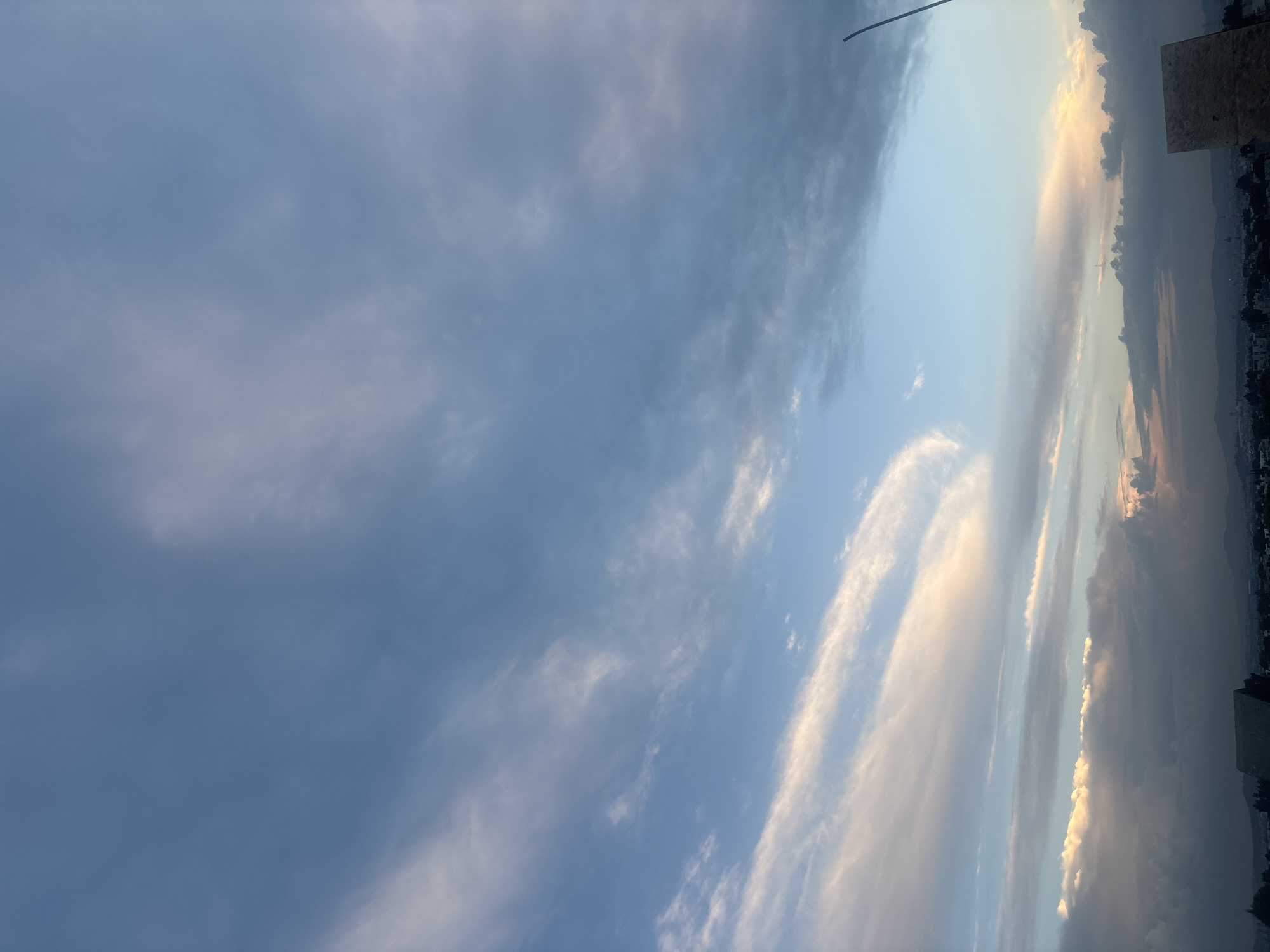 The image captures a sky view in San Pablo del Monte, Mexico. The sky is predominantly blue, filled with scattered clouds. Some clouds show a white to golden hue, likely from the sun. In the rightmost edge of the frame, there are blurred buildings and the dark outline of the mountains.
