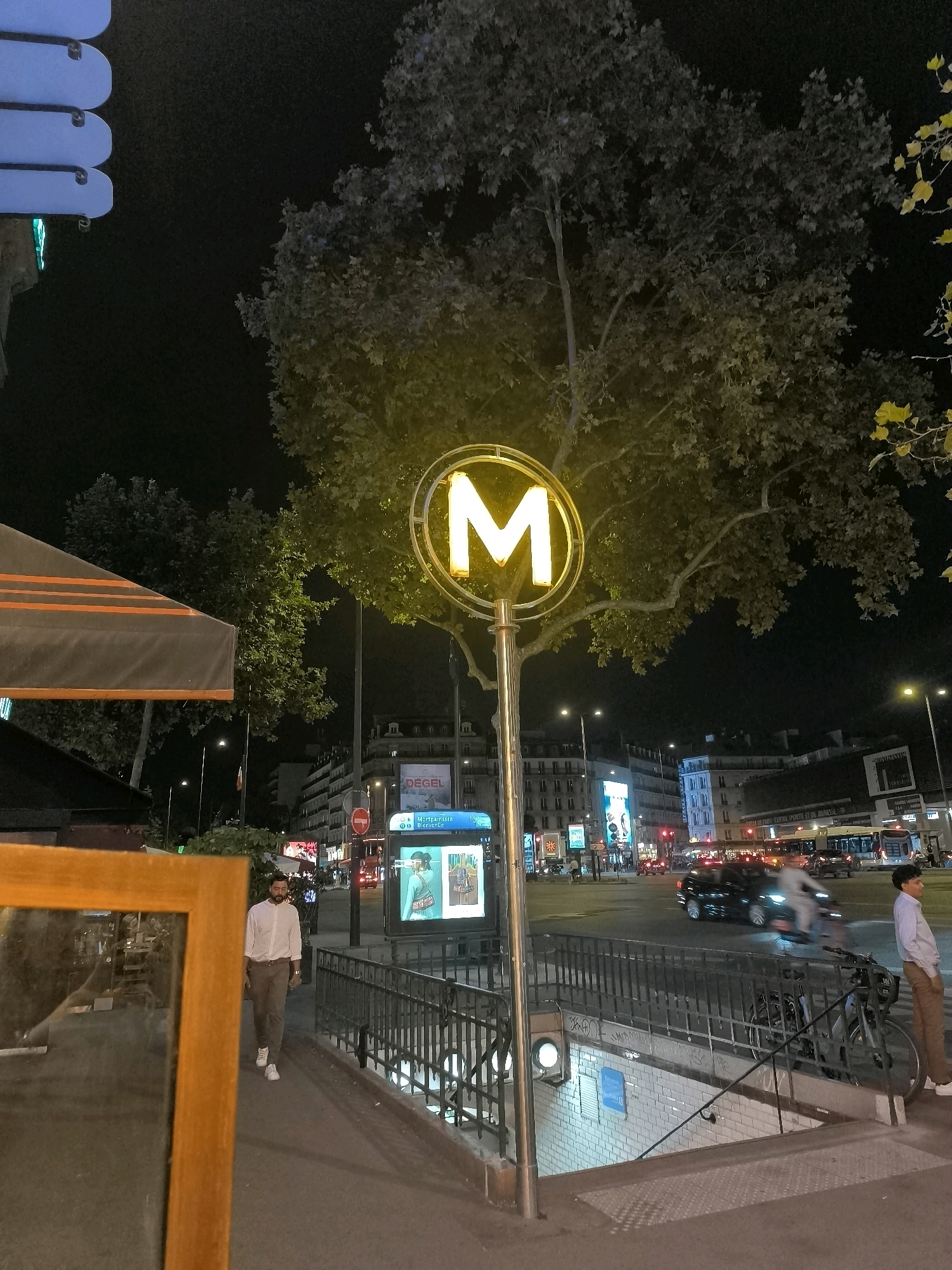 This is a nighttime, eye-level, outdoor photograph taken on a street in Paris, France. A lit "M" sign, characteristic of the Paris Métro, stands prominently in the center of the image, illuminated by a yellow glow. To the left, a portion of a red-and-orange striped awning is visible, suggesting a cafe or outdoor seating area.
A man wearing a light-colored, long-sleeved shirt and dark pants walks along a sidewalk towards the right of the frame. He is wearing white sneakers. A metal railing lines the right side of the sidewalk, separating it from a downward-sloping staircase leading to a Métro entrance. The staircase is illuminated by a blue sign above it. A bicycle is parked next to the railing near the bottom of the stairs.
Across the street, there is traffic, including cars and a bus, with streetlights and building lights creating a vibrant urban nighttime atmosphere. Buildings with multiple windows line the street, some displaying illuminated advertisements. A tree with a full canopy of leaves stands behind the "M" sign, its foliage catching some of the yellow light from the sign. The sky is dark and clear. The overall scene captures a slice of Parisian life at night, centered around a Métro station.