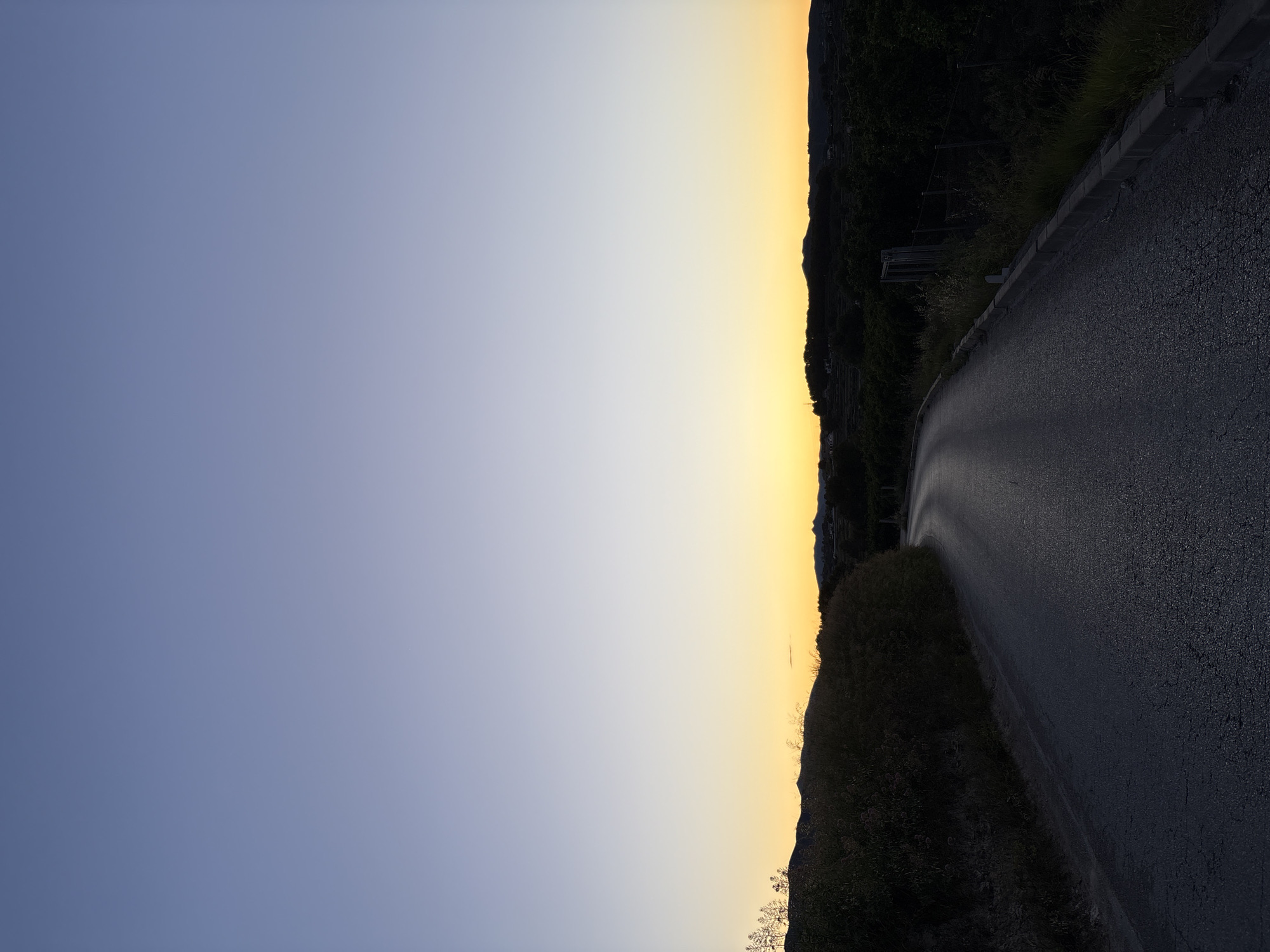 A low-angle, rotated view depicts a road in Teulada, Spain, likely taken during the golden hour. The sky transitions from a deep blue to a golden hue. A paved road curves to the right, bordered by vegetation on both sides. The vegetation includes bushes and taller, darker plants, possibly trees or shrubs along the hillside. A guardrail is visible along the road's edge. The road's surface is asphalt, with a coarse texture.