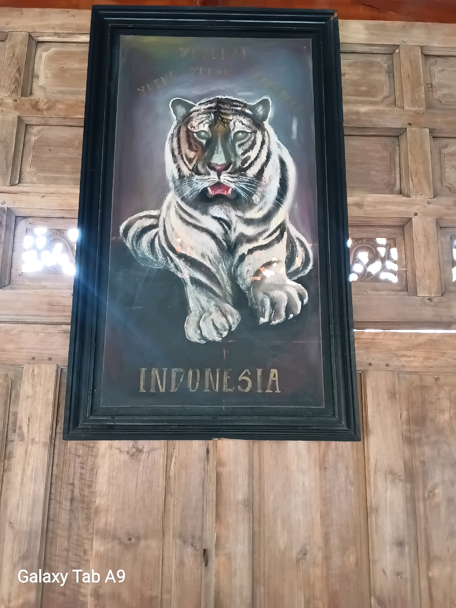 A framed painting of a white tiger with black stripes hangs on a wooden wall. The painting is dark, with the tiger's face and body illuminated. Text is visible above and below the tiger. Above, "YUSUF" is faintly written in white. Below, "INDONESIA" is written in bold, gold letters. The wooden wall appears to be made of vertical planks and has decorative carved square panels on either side of the painting. Natural light streams in from the carved panels on the left, casting a bright glare on the painting. The bottom of the image shows a watermark that reads "Galaxy Tab A9." The image is a photograph of the painting and its surroundings. The style and subject matter suggest it could be a piece of folk art or a patriotic representation from Indonesia.
