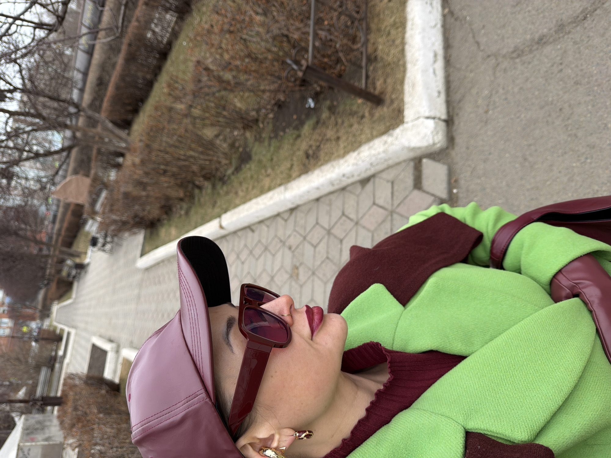 A woman wearing a maroon cap, maroon sunglasses, and a green and maroon outfit is outdoors. She is lying on a paved surface. Trees and bushes are visible in the background, along with a white curb. Buildings are visible in the distance. Based on the characteristics of the trees and architecture in the background, it is possible this image was taken in Krasnoyarsk, Russia.