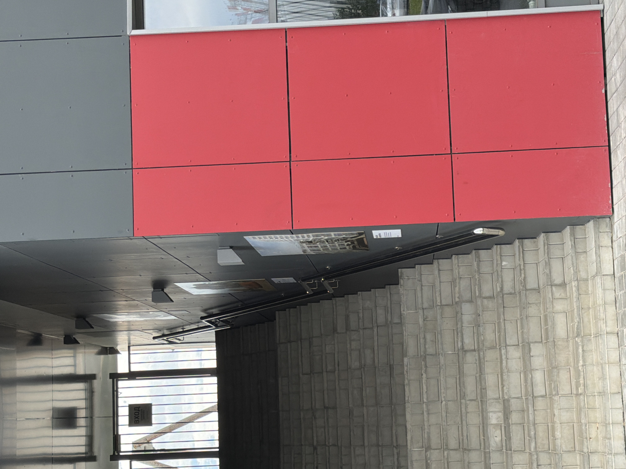 This is a vertical, eye-level, full shot of the exterior of a modern building with a distinctive facade and an outdoor staircase. The building's facade is divided into sections of gray and bright red panels, with a large window at the top. Below the facade, a dark ceiling area is visible, featuring several light fixtures and some posters or signs. To the right of the building, a wide outdoor staircase made of concrete blocks ascends, with a metal railing running alongside it.
In the lower left portion of the image, a portion of a covered walkway or entrance area is visible. This area has a metal door with a sign that reads "ВХОД" in Cyrillic letters, meaning "entrance." The door has horizontal bars across it. Reflections are visible on the metallic surfaces in this area.
The scene appears to be outdoors, possibly during the day, given the natural light. There are no people visible in the frame. The architecture suggests a contemporary or institutional building. The weather appears to be clear. The overall impression is one of a functional and modern urban or commercial environment.