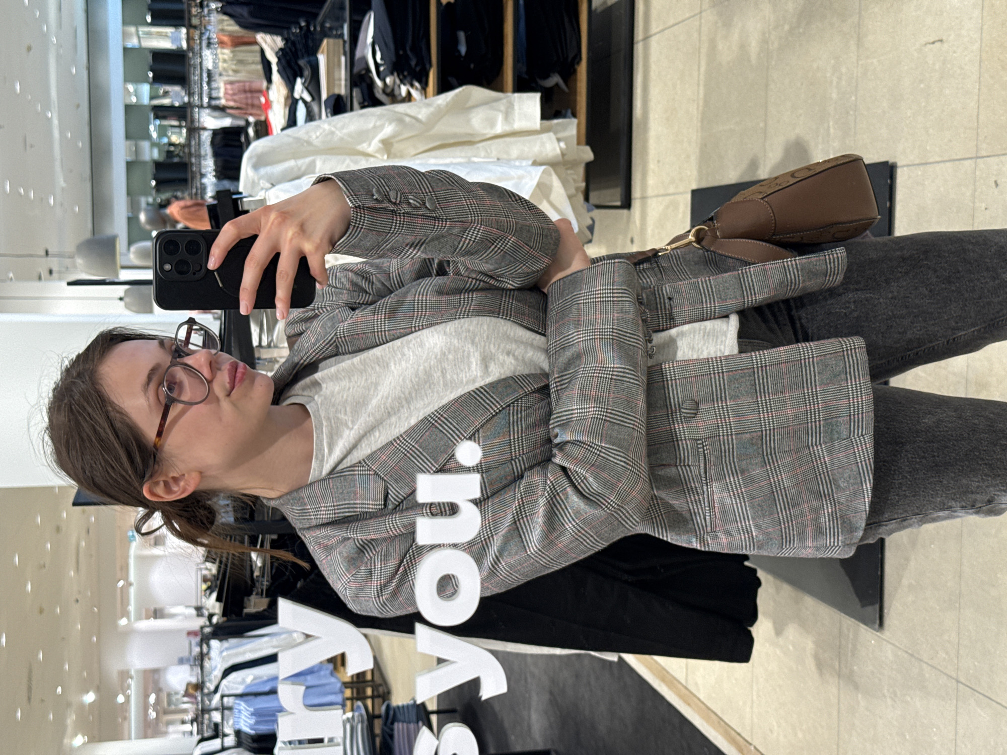 A young woman with brown hair tied back in a ponytail and wearing glasses is taking a selfie with a black smartphone in a clothing store. She is wearing a gray plaid blazer over a white t-shirt and dark jeans. A brown purse with a shoulder strap hangs over her blazer. The store has clothing racks filled with garments visible in the background. A sign with white letters, partially visible, reads "you." The location appears to be an interior setting with tiled flooring. There is no clear indication this is in Budapest, Hungary.