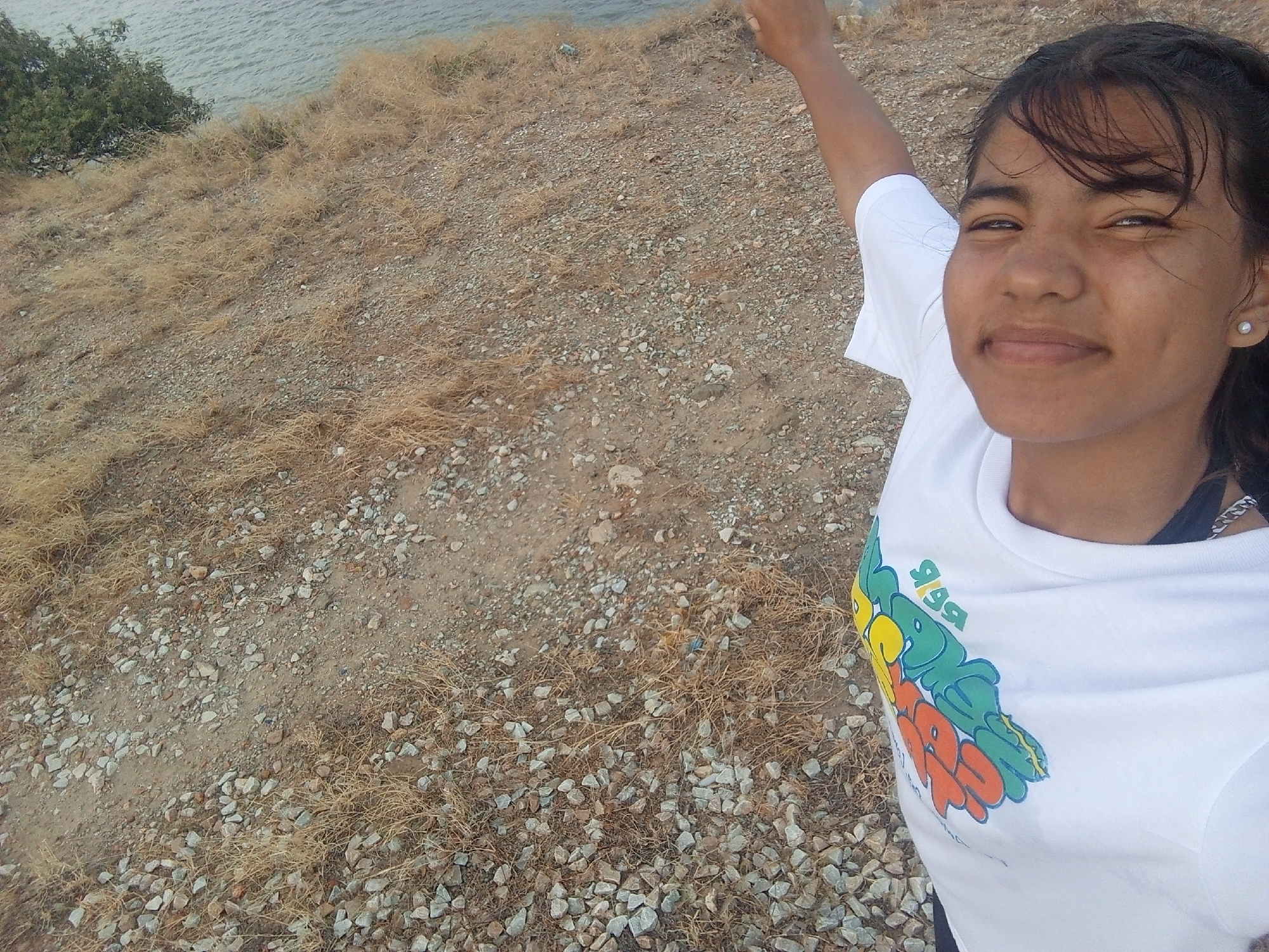 A young woman stands smiling in the foreground. She is light-skinned with brown hair pulled back and wearing a white t-shirt with a colorful logo. In the background, there is a sloping, rocky terrain covered with dry, brown vegetation leading to a body of water. The location is Juan Griego, Venezuela.