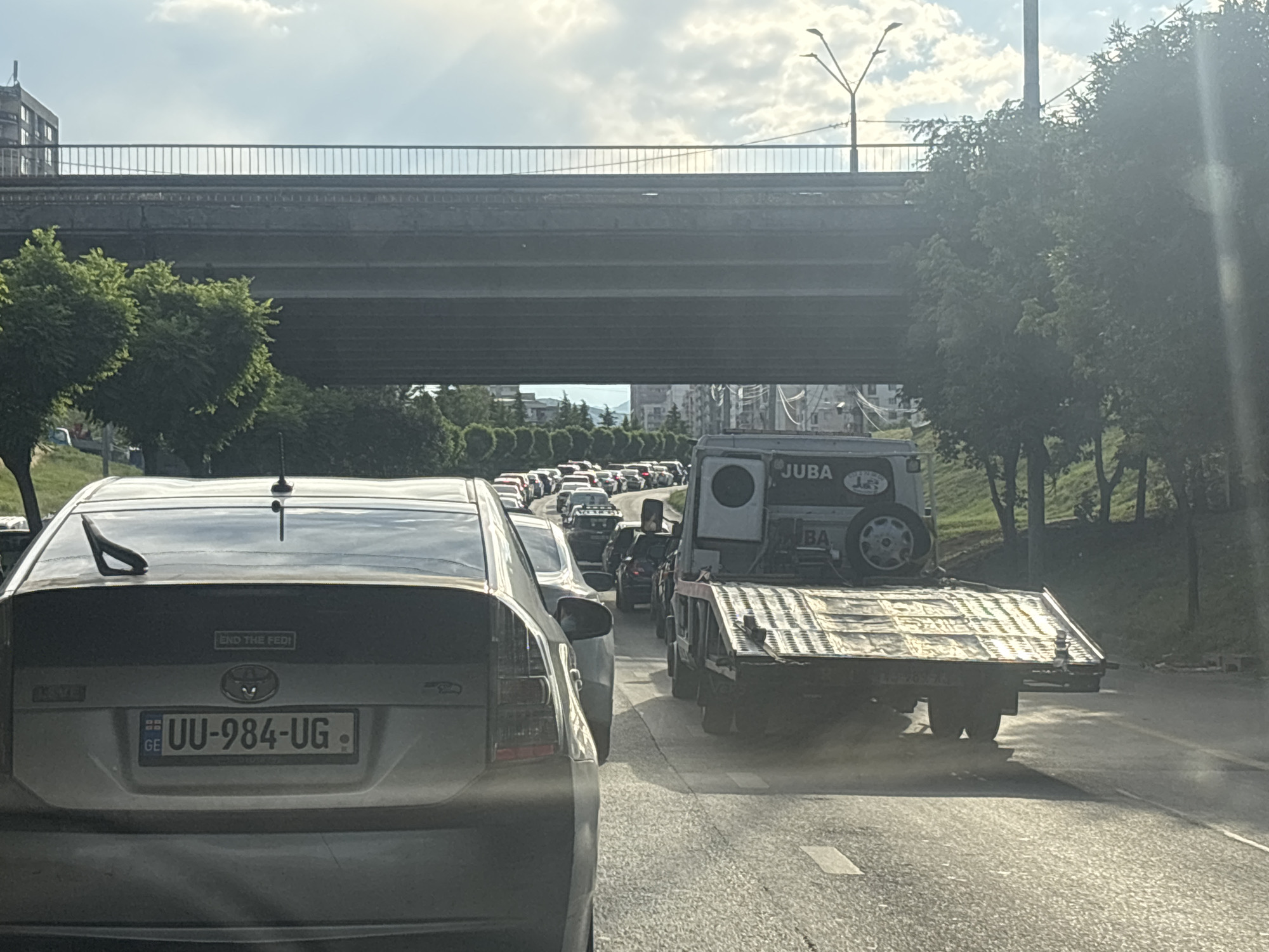 The image captures a traffic jam on a sunny day in Tbilisi, Georgia. A grey Toyota Prius is in the foreground, its rear visible. Behind it, a line of cars stretches into the distance, all stuck in traffic. A flatbed tow truck with "JUBA" written on its side is in the right lane, carrying a vehicle on its platform. The scene is set under a highway overpass, with trees lining the side of the road and buildings visible in the background. The sky is partly cloudy, indicating daytime. A license plate on the Prius reads "UU-984-UG". There is also a sticker on the back of the Prius that says "END THE FED". The overall mood is one of congestion and a typical urban commute.