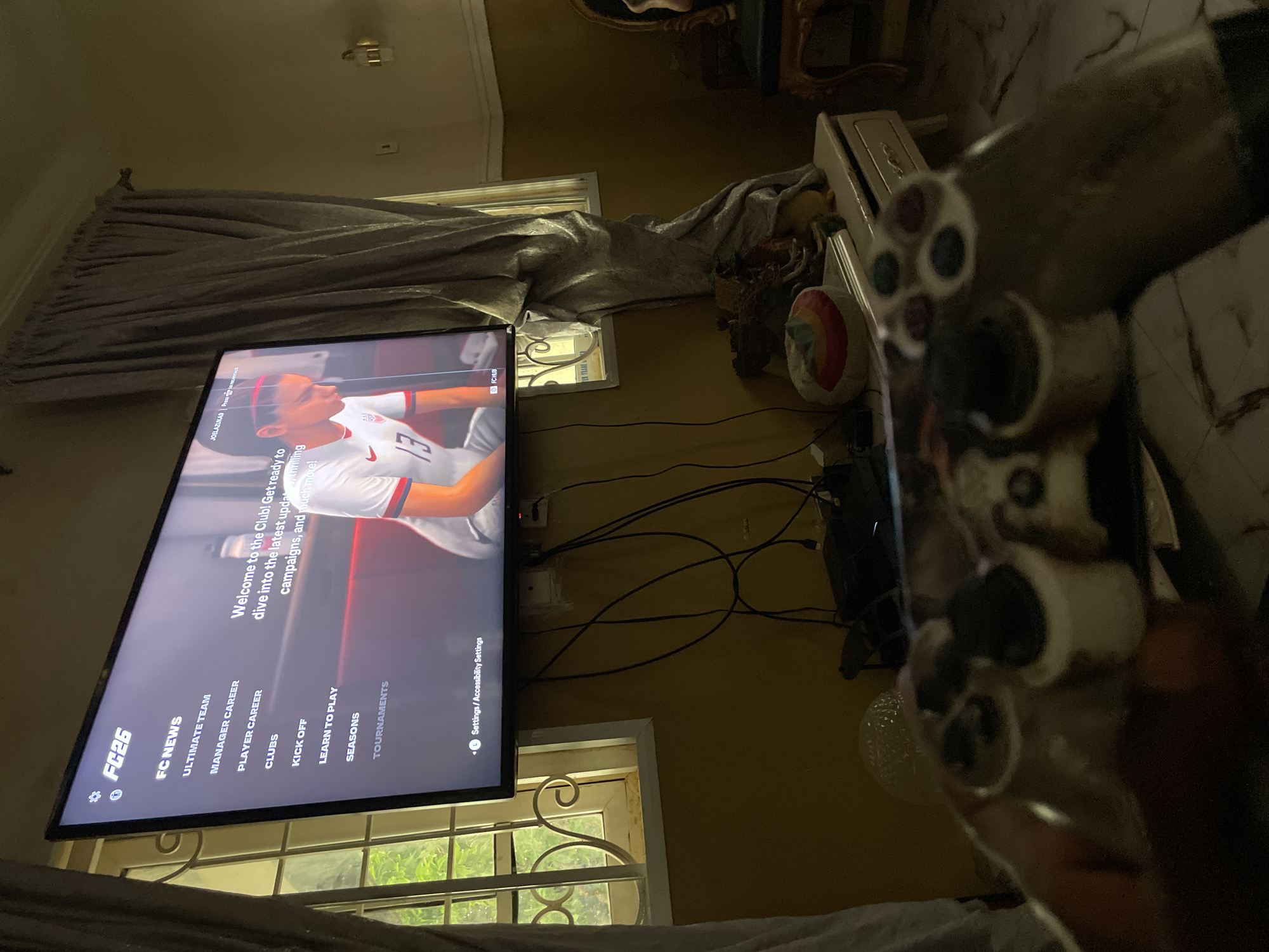 The image shows a person holding a video game controller while sitting in front of a TV screen displaying the FC26 game menu. The TV is mounted on a wall with visible power outlets and cables. Below the TV is a window with metal bars, visible outside. The room is furnished with a table or console. It is unknown whether the location is in the city of Rafin Bauna, Nigeria.
