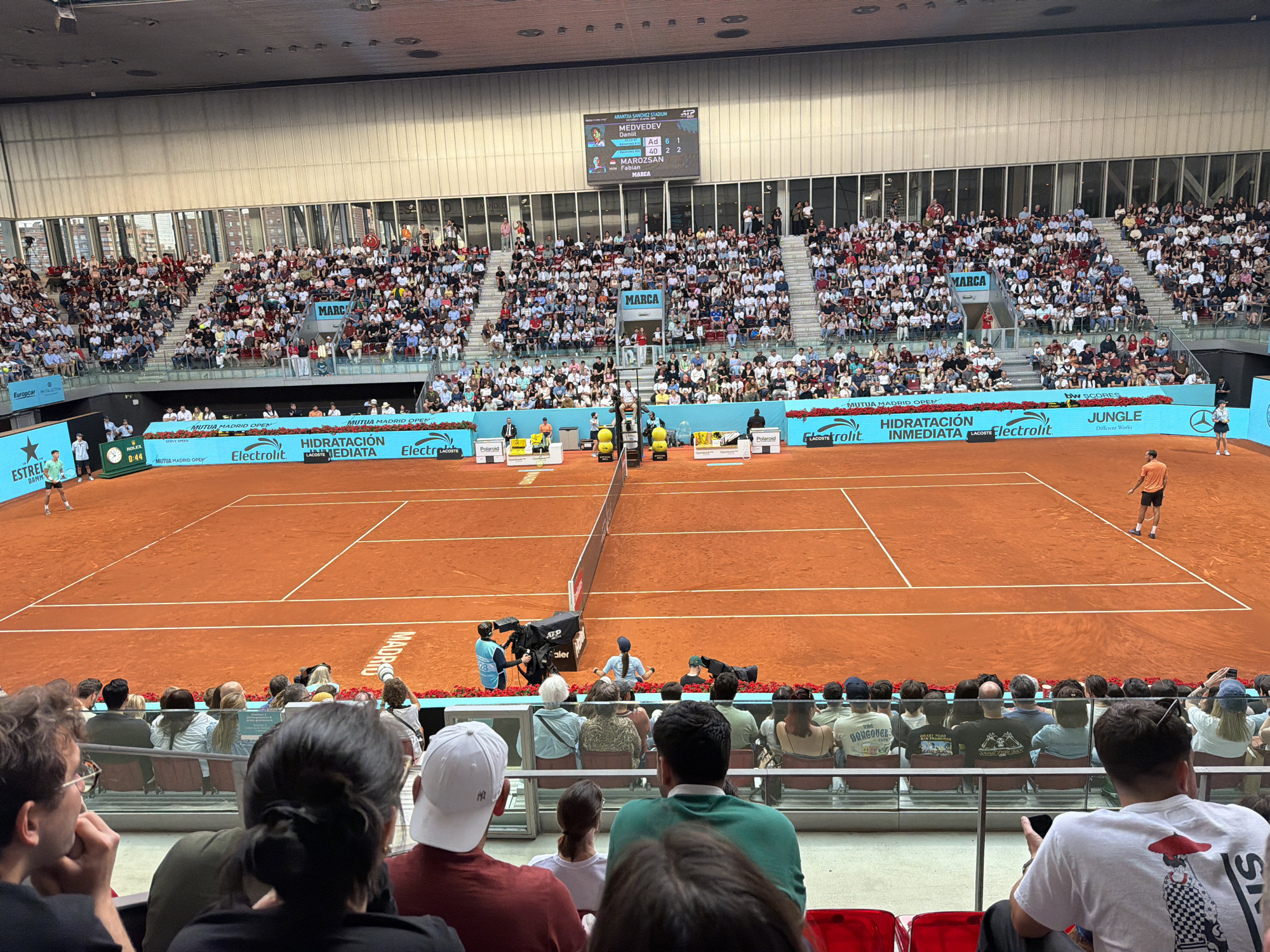 The image shows a tennis match at the Arantxa Sánchez Stadium in Madrid, Spain. The court is clay, and the stands are full of spectators. A scoreboard indicates the match is between Daniil Medvedev and Fabian Marozsan. A camera crew is stationed near the net. Several advertisers' logos are displayed. People are seated in the foreground, watching the match.