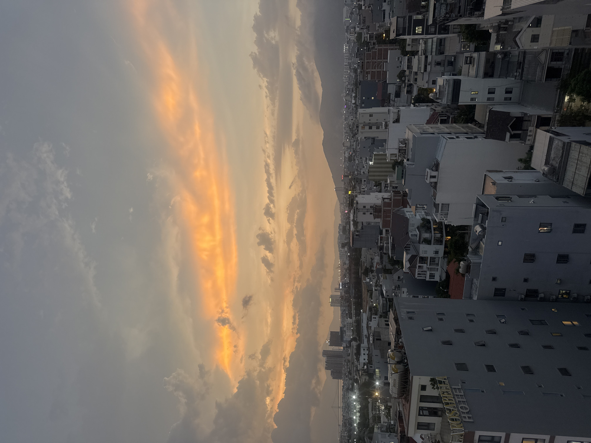 The image shows a view of Đà Nẵng, Vietnam, at dusk or dawn. The sky is filled with clouds, with streaks of orange and yellow light breaking through, suggesting either sunrise or sunset. The city extends along the landscape, with buildings of varying heights and styles. A mountain range is visible in the distance. The buildings appear to be mostly residential, with some commercial establishments. In the foreground, there is a hotel with "ROSABELLA HOTEL" written on it. Streetlights are on, illuminating the streets. There are no people visible in the image.