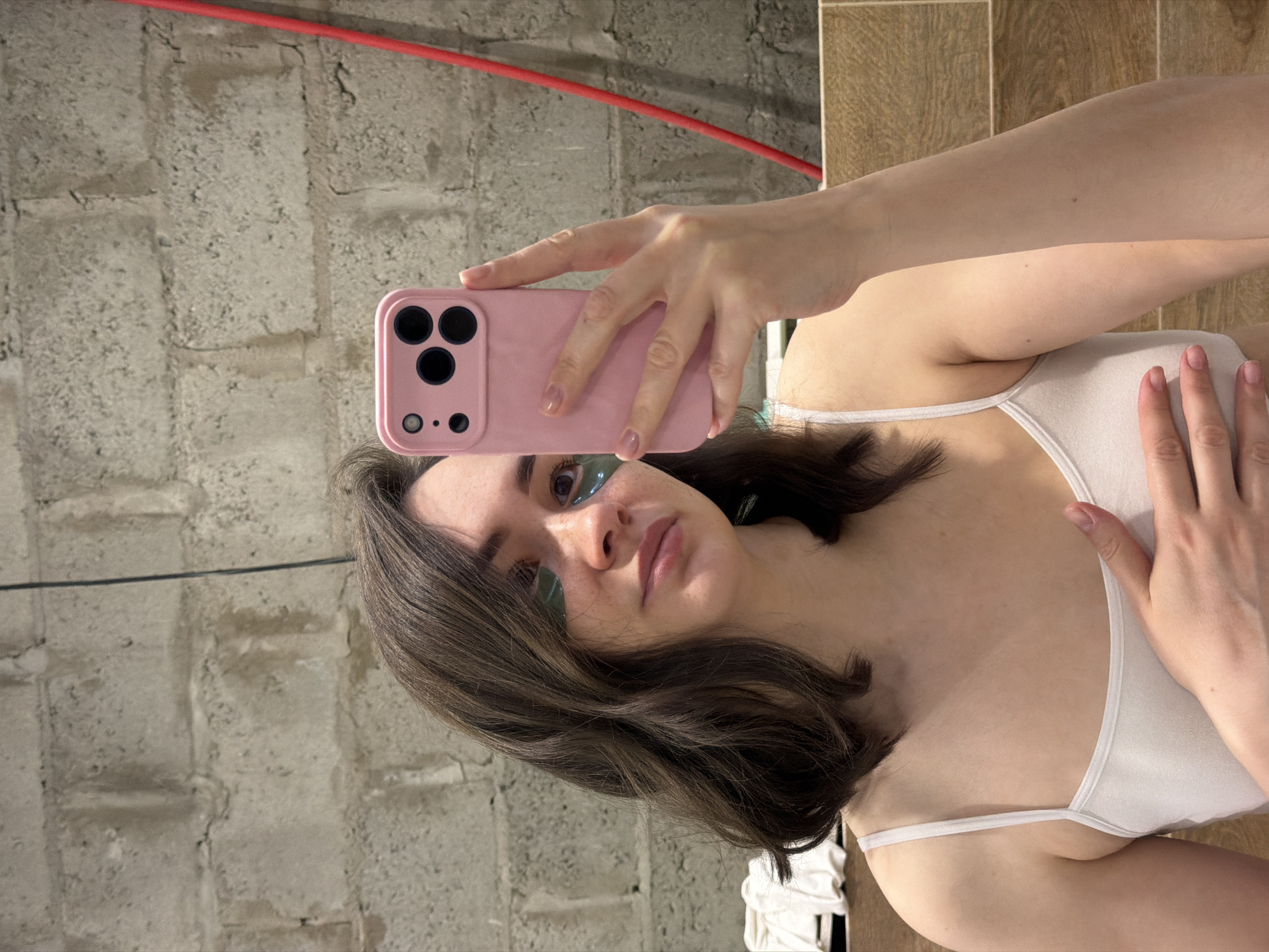 A woman is taking a selfie with a pink smartphone. She is wearing a white tank top and has eye patches on. The background consists of a concrete wall and a wooden surface. The location cannot be determined from this image.