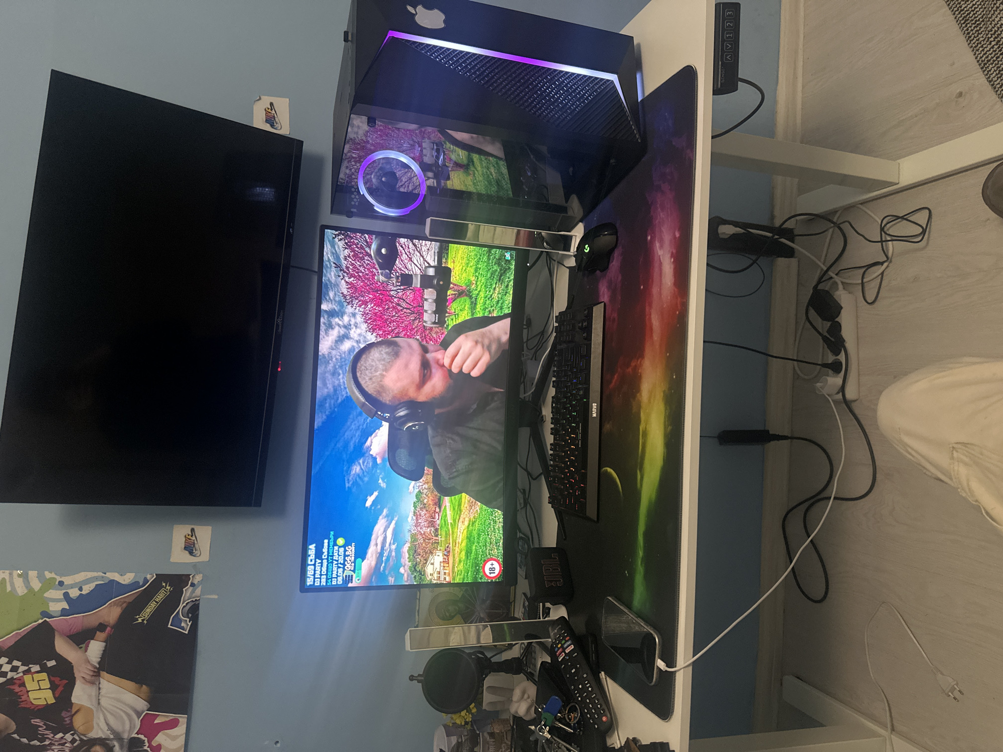 The image captures a detailed indoor scene, likely a gaming or office setup, featuring a white desk against a light blue wall. The primary subject is a computer workstation with a PC tower, monitor, keyboard, and mouse, alongside various peripherals.
The PC tower, black with an Apple logo on top, features internal purple lighting and a visible fan. The monitor displays a vibrant scene of a person wearing headphones, set against a backdrop of green fields and pink-blossomed trees. A red "18+" age rating icon is visible on the screen. To the left of the gaming setup, a large black flat-screen TV is mounted on the wall.
On the white desk, a colorful, galaxy-themed mouse pad covers a significant area. Other items include a black mechanical keyboard with "MARVO" branding, a black gaming mouse with a green "G" logo (Logitech), a microphone on an arm, a soundbar, a "JBL" brand portable speaker, a smartphone, multiple remote controls, and a set of keys. Under the desk, cables are visibly managed (or unmanaged) with power strips. A desk controller with "EZDAV" branding and buttons "1 2 3" and up/down arrows is attached to the desk.
On the light blue wall, a small "NIKE" sticker is placed above the large TV. Below and to the left of the TV, a poster is partially visible, showing a person and the text "SUNDAY HABIT".
Visible text on the monitor includes "15/69 СЪБА", "DJ PARTY", "283 Общо Събова", "54 ОБЩО УТ МЕМБЪРИ", "DJ PARTY ДАТИ", and "06.06/20.06". The overall impression is a personal space dedicated to computing, entertainment, or content creation.