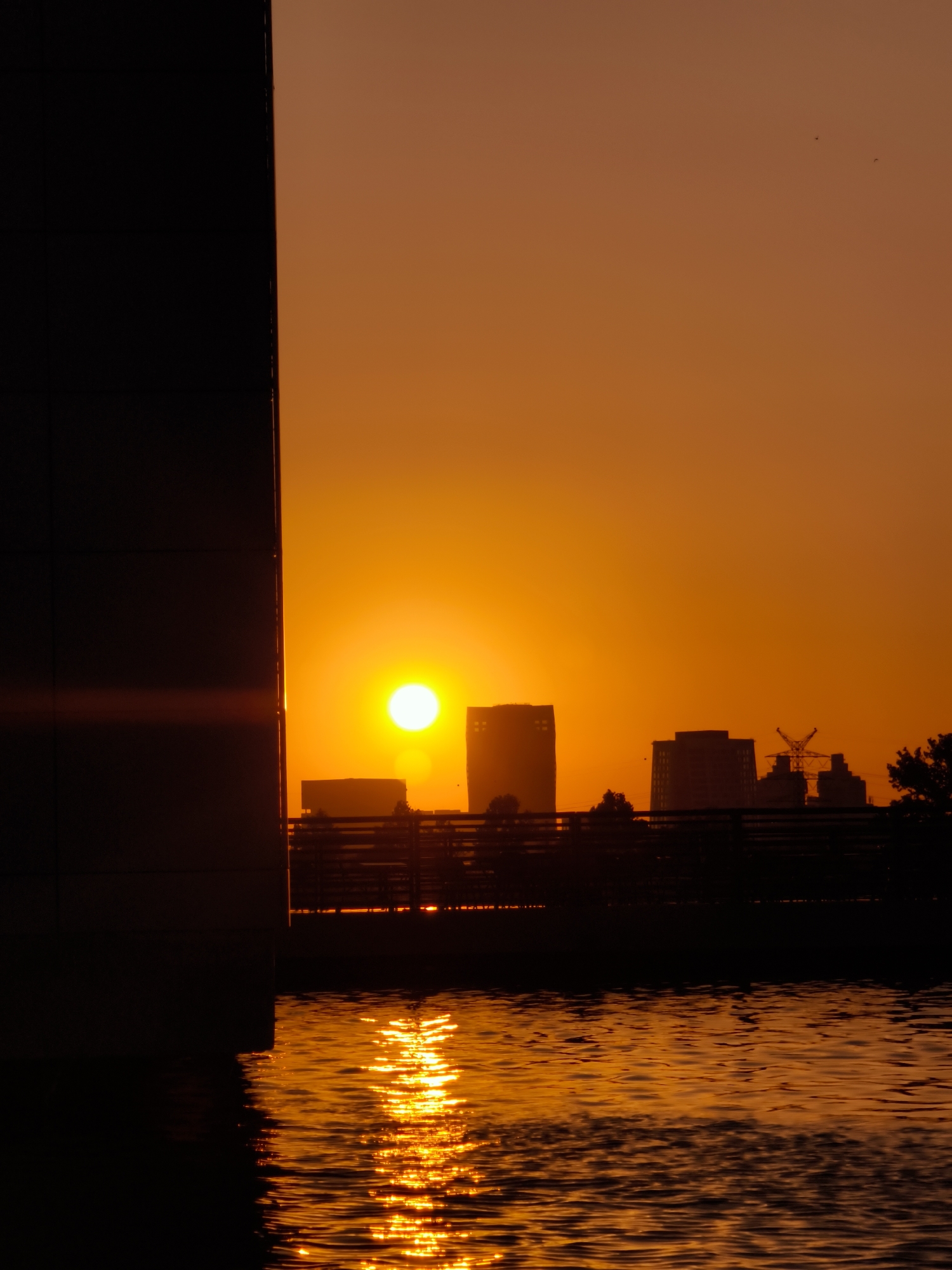 A vertical, full shot captures the vibrant orange sunset over the Be'er Sheva skyline, reflected in the gentle ripples of a body of water. The sun, a bright, dominant circle, hangs low in the sky, casting a warm glow that saturates the entire scene with hues of orange and yellow. To the left, a large, dark, rectangular structure, possibly a building or a wall, dominates the foreground, its silhouette stark against the luminous sky.

In the mid-ground, a silhouetted cityscape emerges. Several buildings of varying heights form a distinct line against the horizon. The most prominent are two taller, rectangular structures, one appearing to be a skyscraper with a flat top, and another slightly shorter building next to it. To the right of these, a distinctive lattice tower, resembling a construction crane or a communication tower, stands out against the sky. A dark, horizontal line, likely a fence or railing, runs across the mid-ground, separating the water from the buildings and creating a sense of depth.

The water in the foreground is a mirror to the sky, reflecting the intense orange of the sunset with a shimmering, almost liquid gold effect. The sun's reflection creates a bright, unbroken path across the water's surface, highlighting the texture of the ripples. A few faint, dark specks, possibly birds, are visible in the upper part of the sky, adding a subtle sense of movement to the otherwise still scene. The overall atmosphere is one of serene beauty, with the natural spectacle of the sunset juxtaposed against the urban landscape of Be'er Sheva.