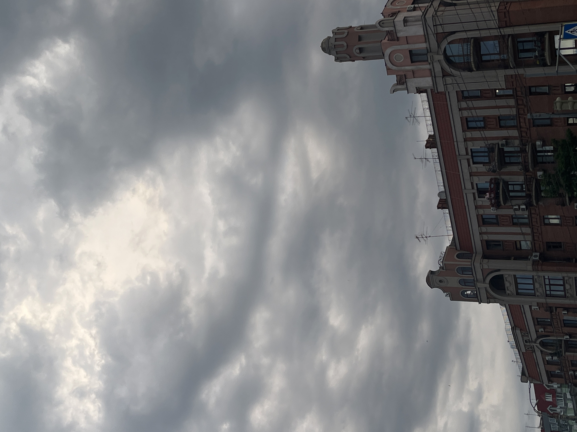 The image is a vertical, low-angle shot of a cloudy sky and a multi-story building, oriented such that the building appears to be growing out of the sky. The building is reddish-brown with lighter pinkish-beige architectural details, including arched windows and decorative elements at the top. Several windows are visible, some with air conditioning units below them. Balconies with railings are also present on some floors. A traffic sign is partially visible on the right side of the frame. Several antennas are mounted on the roofline of the building. The sky is filled with gray and white clouds, with some brighter patches suggesting daylight, though the overall mood is overcast, possibly indicating approaching rain or a gloomy day. The image is likely taken in Samara, Russia, given the location context. No people are visible in the image.