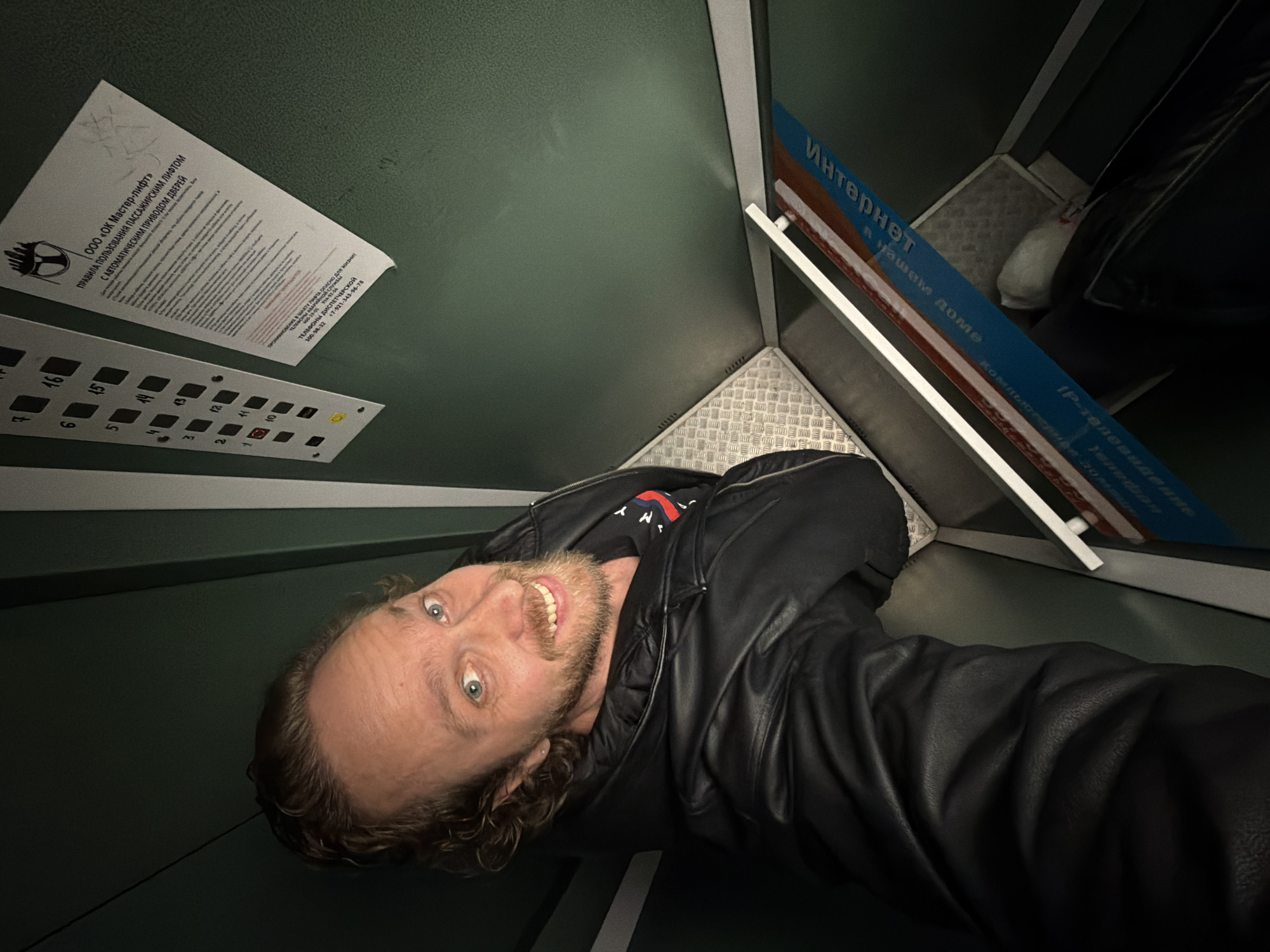 A man is taking a selfie inside an elevator. He has fair skin, blue eyes, and light brown hair. He is wearing a black leather jacket over a black t-shirt. The elevator has green walls and a textured metal floor. There is a button panel with floor numbers. A sign with the text "ООО «ОК Мастер-лифт» ПРАВИЛА ПОЛЬЗОВАНИЯ ПАССАЖИРСКИМ ЛИФТОМ С АВТОМАТИЧЕСКИМ ПРИВОДОМ ДВЕРЕЙ" is attached to the wall. Another sign states "Интернет в нашем доме" and includes references to computers and IP television. The man is likely in an elevator in Saint Petersburg, Russia, based on the Cyrillic script and the phone number listed on the elevator rules sign.