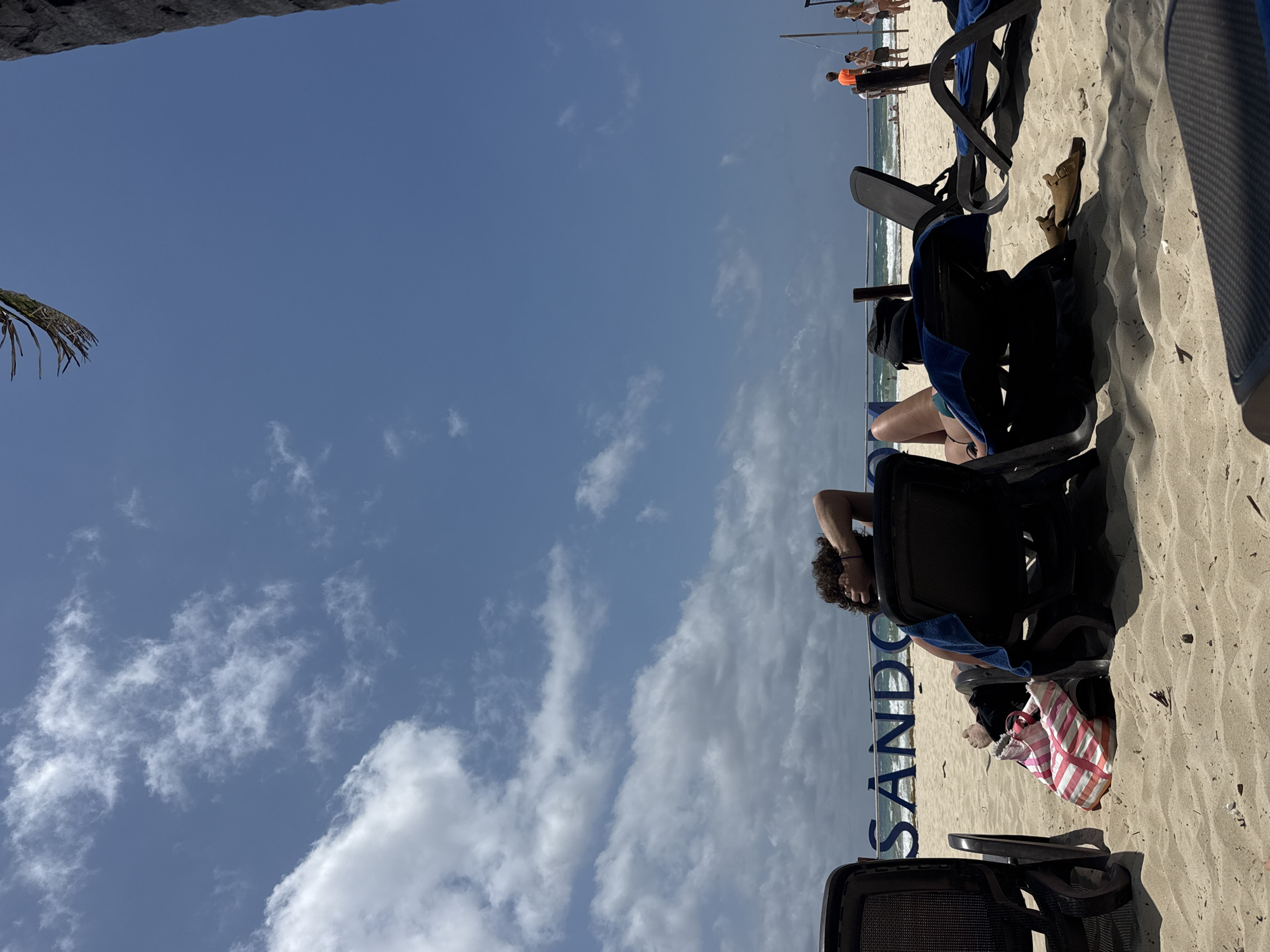 The image shows a beach scene in Playa del Carmen, Mexico, possibly at the Sando hotel or resort as the word "Sando" is visible. There are several beach chairs, some occupied by people relaxing. One person is visible in a chair, possibly sunbathing, with a striped bag nearby. The beach leads to the ocean, with a few other people visible in the distance near the water. The sky is blue with scattered clouds. The scene is photographed from a low angle, looking upwards towards the sky.