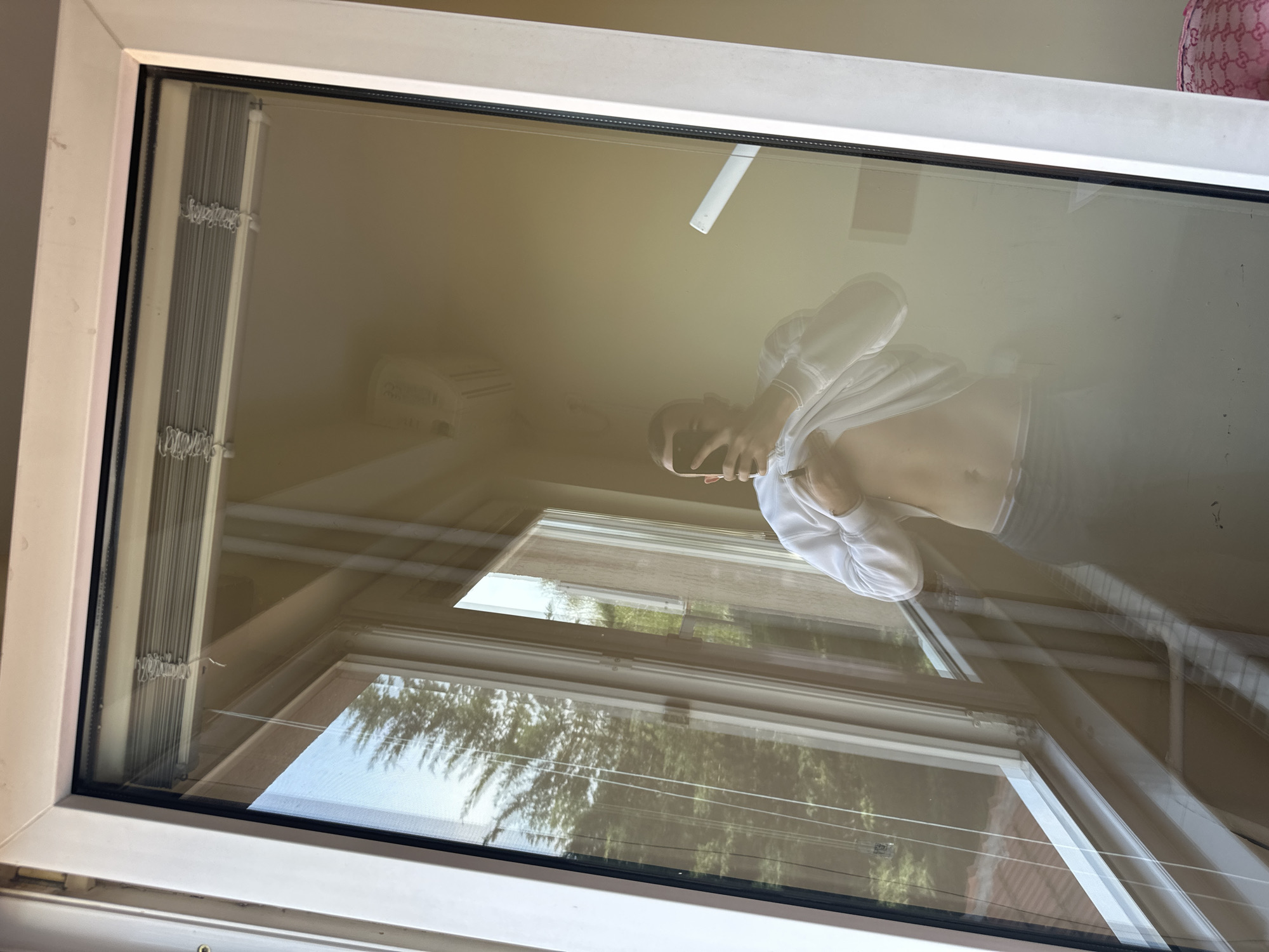 The image is a view through a large window, dominated by a strong reflection of the interior space and the person taking the photograph. The window itself has a white frame and features internal, accordion-style blinds visible along the left edge, with text that appears to read "Symphony pleated".
A person is clearly reflected in the glass, holding a black smartphone to capture the image. They are wearing a white long-sleeved top, which is pulled up to expose their midriff and upper abdomen. Dark bottoms, possibly jeans, are also visible.
Beyond the reflection, an outdoor scene can be seen through the glass, showing green trees and what looks like a building with a reddish-tiled roof, suggesting it is daytime.
Further details are visible in the reflections within the room. In the upper right corner, a pink item, potentially a bag or piece of clothing, displays a repeating pattern that strongly resembles the "Gucci" brand logo. A faint outline of what might be an air conditioning unit or a light fixture is also discernible in the upper left of the reflection. The photo is tilted, giving the window frame a diagonal orientation. The setting is Plovdiv, Bulgaria.