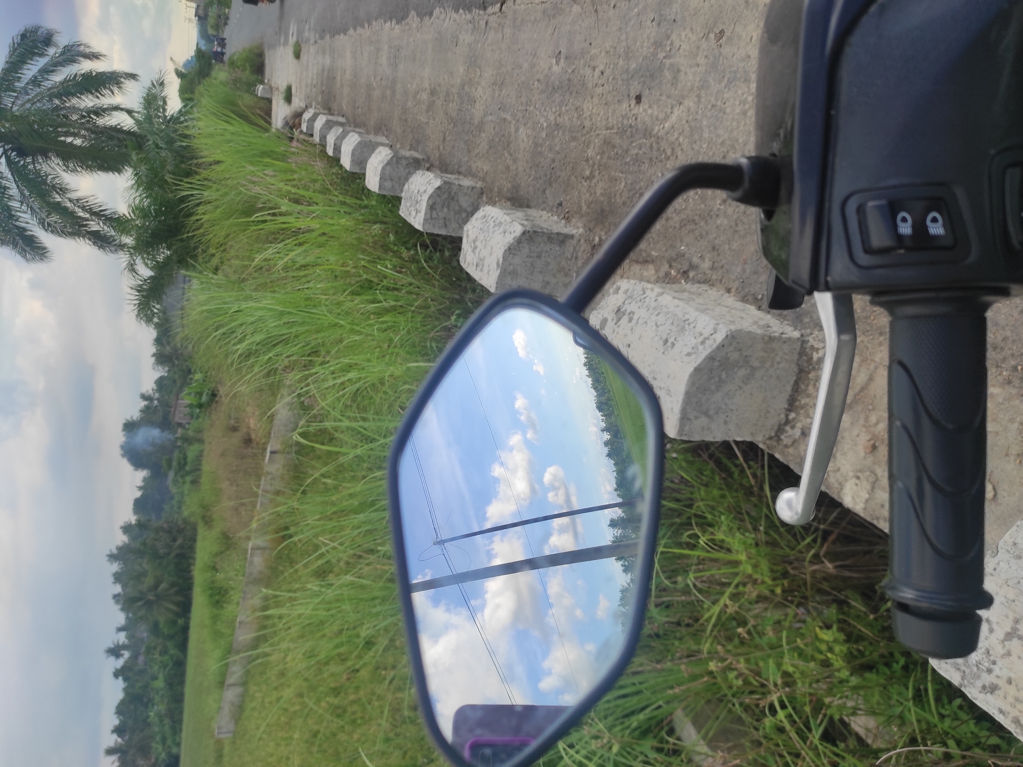 The image presents a view from a motorcycle's perspective in Lhokseumawe, Indonesia, showing the landscape and a reflection in the side mirror. The foreground prominently features the motorcycle's handlebar, grip, and the side mirror, which reflects the sky with clouds and power lines. The background showcases a roadside scene: a concrete curb and sidewalk, green grass growing along the edge, and trees including a palm tree in the distance. The sky is visible in the background, suggesting an overcast day. The vegetation appears lush, indicating a tropical climate. There are no people visible in the primary view, but a distant figure can be seen on the road.