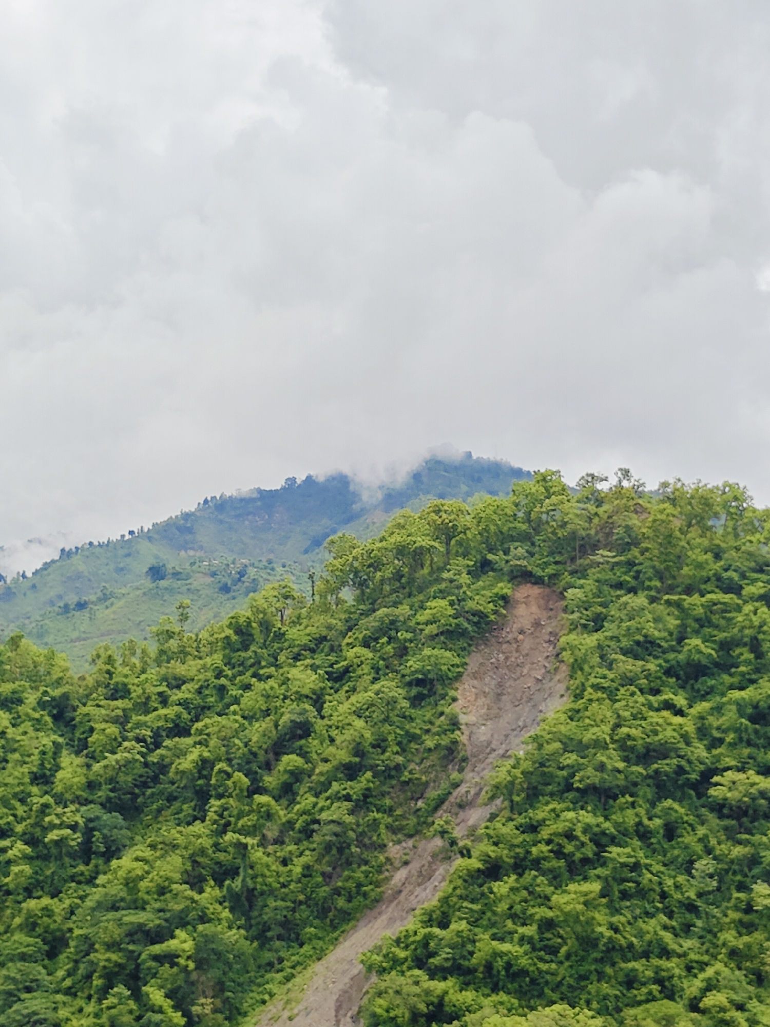 A medium shot captures a landslide scar on a densely forested mountainside in Ichchhakamana, Nepal. The scar, a bare patch of earth, slopes diagonally from the upper right to the lower left of the frame. It is surrounded by a vibrant green forest that covers the rest of the mountain. The sky above is overcast with thick, grey clouds. In the distance, the peaks of other mountains are visible, some shrouded in mist, with hints of vegetation and scattered structures suggesting human habitation. The overall scene depicts a natural landscape shaped by both lush growth and geological events.