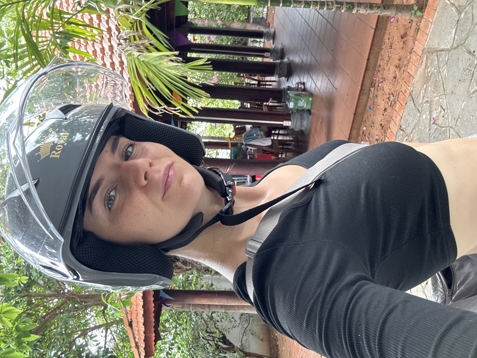 The image features a young woman wearing a black motorcycle helmet with a clear visor. The helmet prominently displays the brand name "Royal" alongside a crown logo on its side. She has light skin, light-colored eyes, and visible freckles across her nose and cheeks, looking calmly towards the camera with a subtle, gentle smile. She is dressed in a form-fitting black ribbed crop top.
The setting is an outdoor, tropical environment, rich with lush green foliage, including prominent palm leaves in the foreground and background. Behind the woman, there are wooden pillars and structural elements that suggest an open-air building, possibly a cafe or restaurant, with visible tables and chairs. The ground transitions from terracotta-colored paving tiles to grey stone pavers, with a strip of reddish soil and small pink flowers visible between them.
The bright, natural lighting indicates it is daytime, likely a sunny morning or afternoon. The presence of the helmet suggests the woman is either about to embark on or has recently completed a motorcycle journey. The tropical plants and architectural style are consistent with the provided location context of Mũi Né, Vietnam.