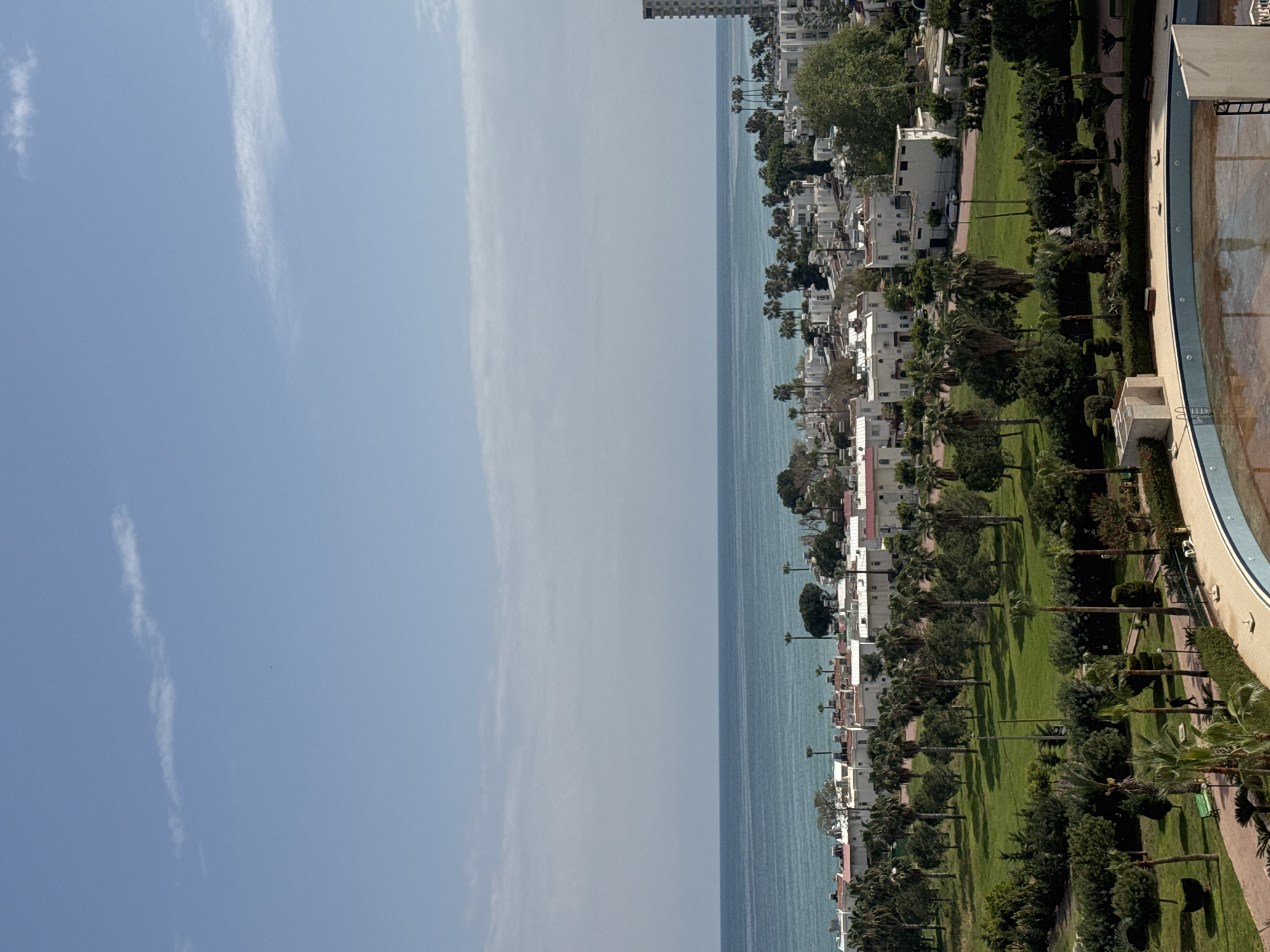 The image depicts a coastal scene in Erdemli, Turkey, featuring a clear blue sky with wispy clouds above the turquoise Mediterranean Sea. A row of white buildings lines the coastline, interspersed with palm trees and green vegetation. A green park area is visible, separating the buildings from a partially visible swimming pool. The overall scene portrays a serene coastal environment.