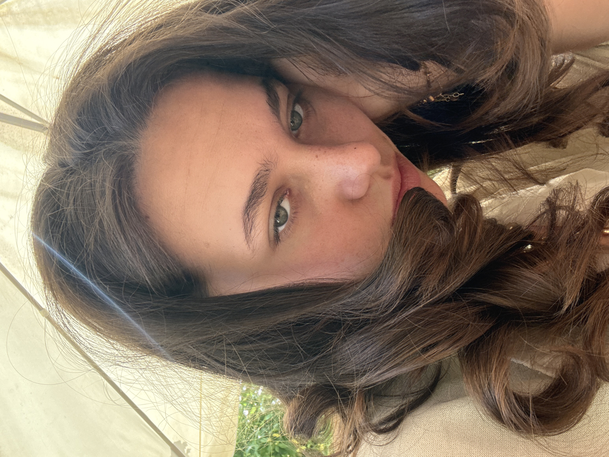 This media file is a close-up, vertically oriented selfie of a young woman. She has long, dark brown wavy hair that frames her face, and light green eyes. Her head is tilted, and she is looking directly at the camera with a neutral expression. Freckles are visible on her nose and cheeks. She is wearing a light-colored garment, possibly a shirt or dress, with a delicate gold necklace visible.
The background suggests an outdoor setting. A blurred green landscape with foliage is visible in the lower portion of the image, indicating it might be daytime. A white or light-colored fabric structure, possibly a tent or canopy, is also present, casting a shadow and creating some diffused light. The lighting suggests it could be a sunny day, possibly late morning or afternoon, with the sun creating a slight glare on the left side of the frame. The overall mood is relaxed and informal. No visible text is present in the image.