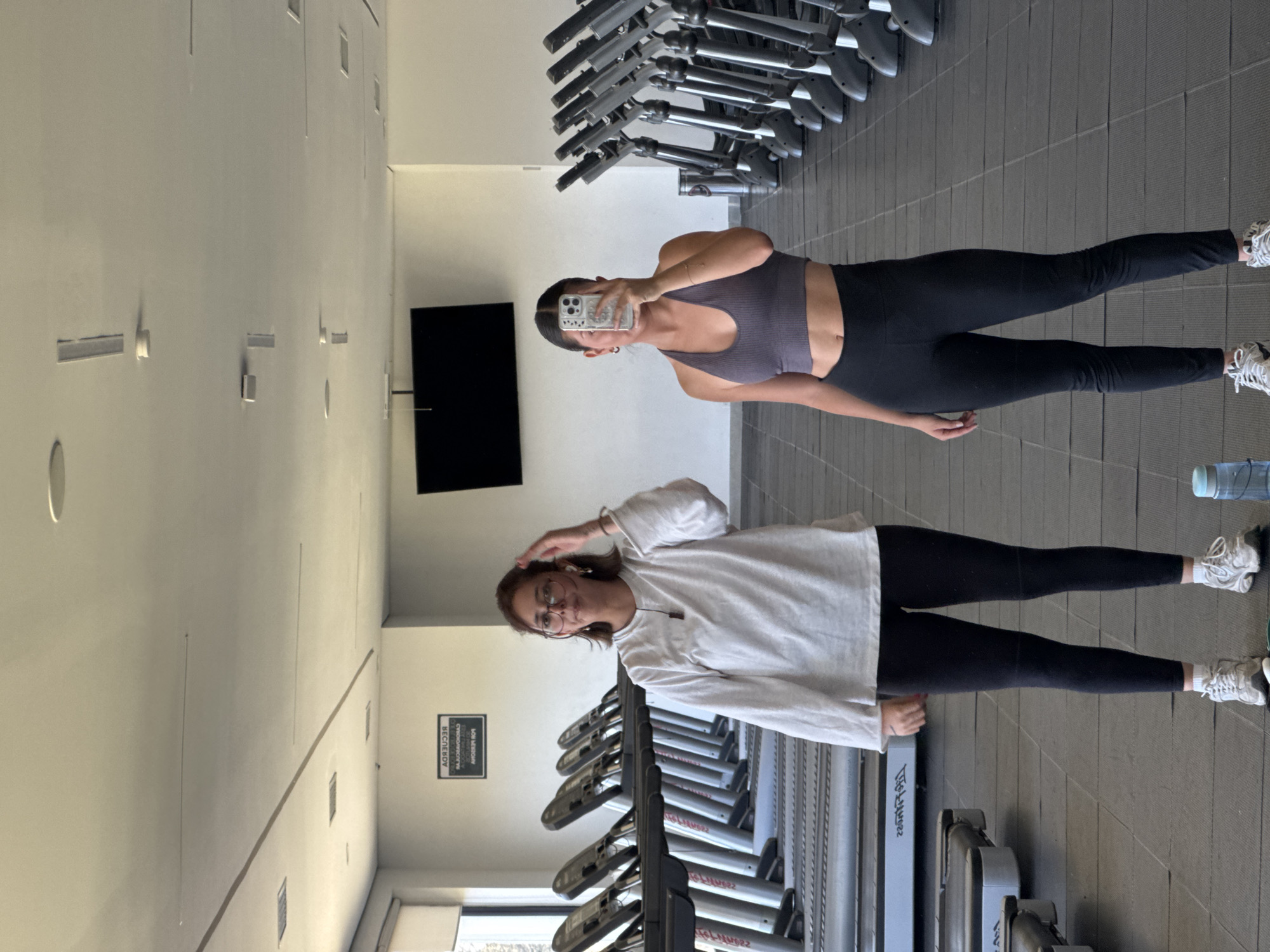 A brightly lit gym in San Andrés Cholula, Mexico, serves as the setting for this image, which captures two women amidst rows of treadmills. The photo appears to be a selfie, as one woman is actively holding a smartphone, reflecting her face and the device.
The woman on the right wears a form-fitting dark purple or grey sports bra and black leggings, paired with white athletic shoes. Her dark hair is pulled back, and she holds the phone up, partially obscuring her face as she takes the picture. To her left, the second woman stands in a relaxed pose. She is dressed in a loose white long-sleeved shirt, black leggings, and white sneakers, with glasses framing her face and light brown hair. Her hand is raised casually near her head, and she offers a slight smile towards the camera.
The gym environment features a dark grey tiled floor, likely rubber, and light-colored walls and ceiling. Numerous treadmills with grey and silver accents line the background, indicating a well-equipped fitness facility. A large black flat-screen television is mounted on a wall. A blue water bottle is visible on the floor near the second woman's feet. A white sign on the wall, appearing mirrored in the reflection, displays the text: "RECUERDA CUIDAR EL MEDIO AMBIENTE" (Remember to take care of the environment) and below it, "BE RESPONSIBLE". The scene suggests a casual moment taken during a visit to the gym.