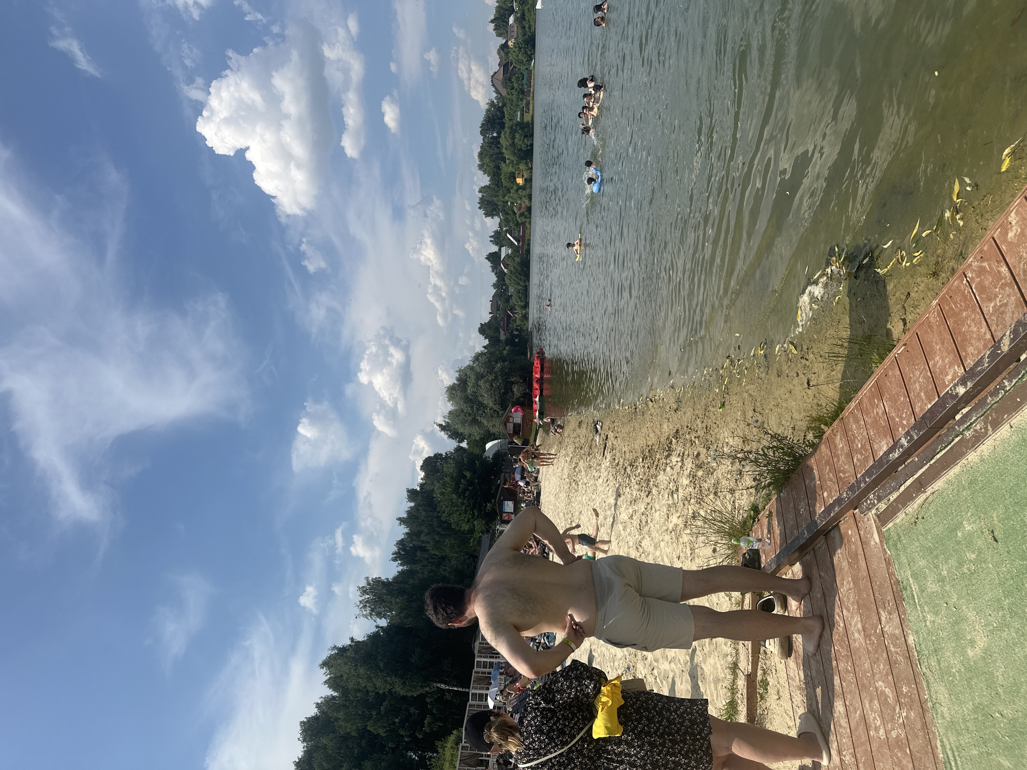 A sunny day at a lake beach in Molokovo, Russia, with a sandy shore, wooden pier, and a body of water filled with swimmers. The sky is blue with scattered white clouds. On the beach, several people are visible, some sunbathing and others walking. Two individuals, a woman with a black and floral dress and a yellow purse, and a shirtless man wearing light-colored shorts, stand on a wooden pier in the foreground, facing the lake. The man has his hands on his hips. The lake is populated with many people swimming, including some with inflatable rings. In the background, trees and houses are visible along the shoreline. Red inflatable watercraft are also present on the water.