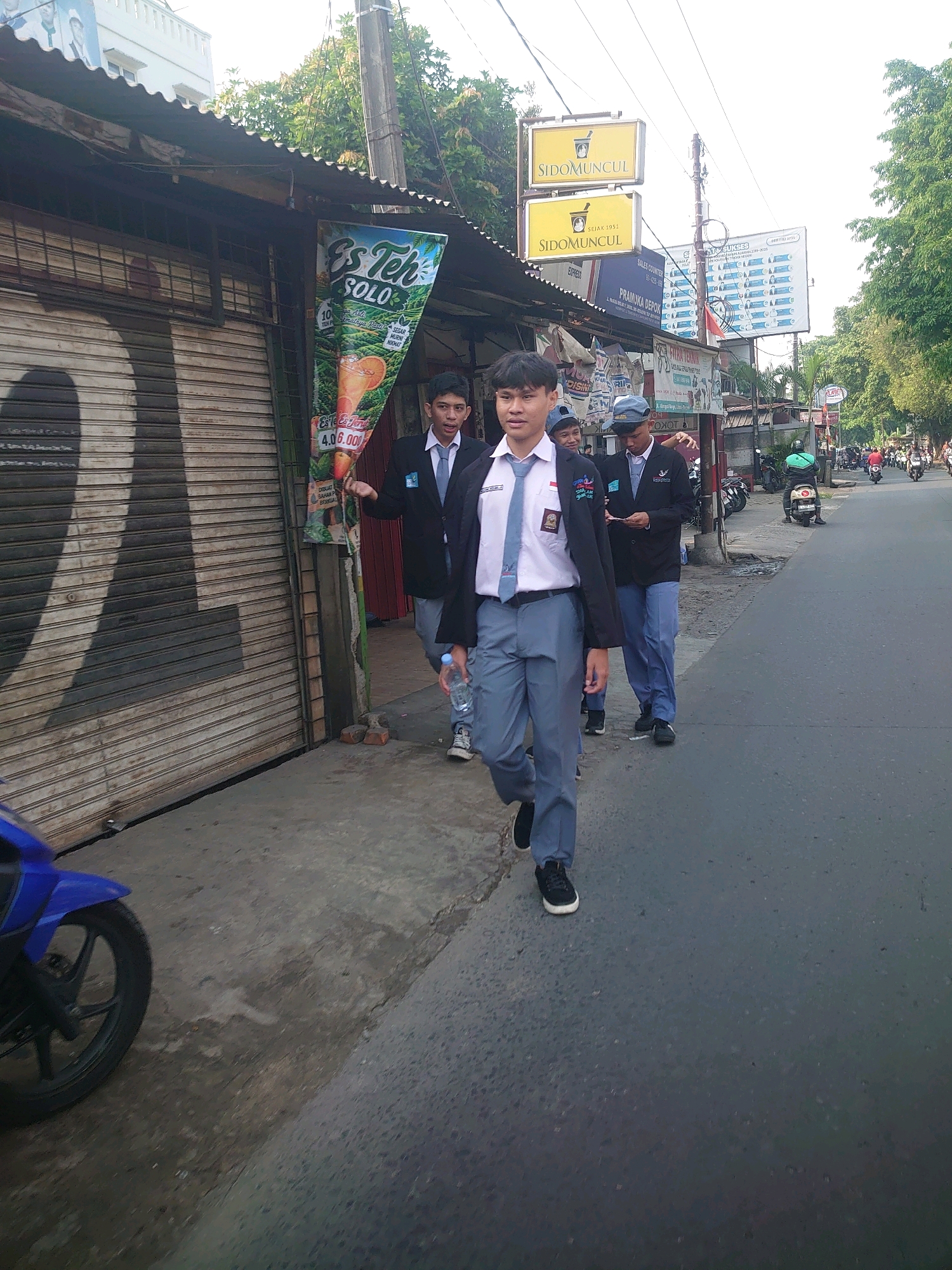 In Depok, Indonesia, several male students in school uniforms are walking along a sidewalk next to a street. The students, dressed in black blazers, white shirts, blue ties, and gray pants, are walking towards the viewer. The student in the foreground is holding a clear plastic bottle. Behind them, a blue motorcycle is parked on the left side of the sidewalk. The sidewalk is adjacent to a street with moderate traffic, including motorcycles and cars. Buildings line the street, with one building featuring a corrugated metal roof and large, roll-down metal doors. Several signs are visible, including advertisements for "SIDOMUNCUL" and "Es Teh SOLO." The environment appears to be an urban commercial area with dense vegetation and trees visible in the background. The lighting suggests it is daytime.