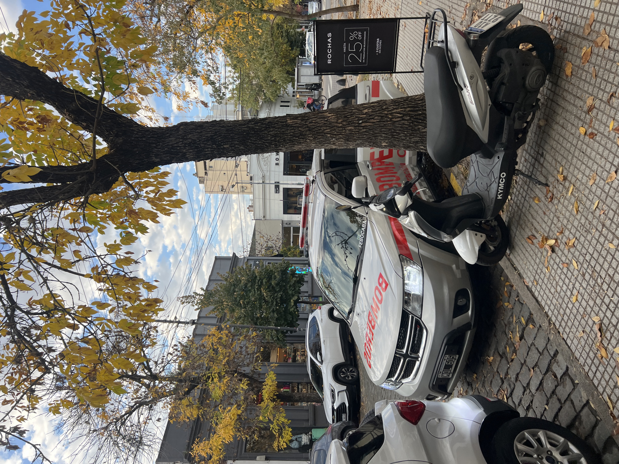 The scene is an urban streetscape in Buenos Aires, Argentina. A tree with yellow leaves dominates the left side of the frame. Beside the tree and parked on the cobblestone sidewalk is a Kymco scooter. Next to the scooter is a light-colored Dodge Journey SUV, clearly marked as a "BOMBEROS FEBOCABA" vehicle - i.e. belonging to the Buenos Aires Fire Department. The text "BOMBEROS" is prominently displayed in red on the hood. A sign for "ROCHAS" offers "HASTA 25% OFF" ("Up to 25% off") on selected products. Several other vehicles, including a white hatchback, are parked along the street. The buildings visible in the background suggest a commercial or residential area. Power lines are visible against a cloudy sky.
