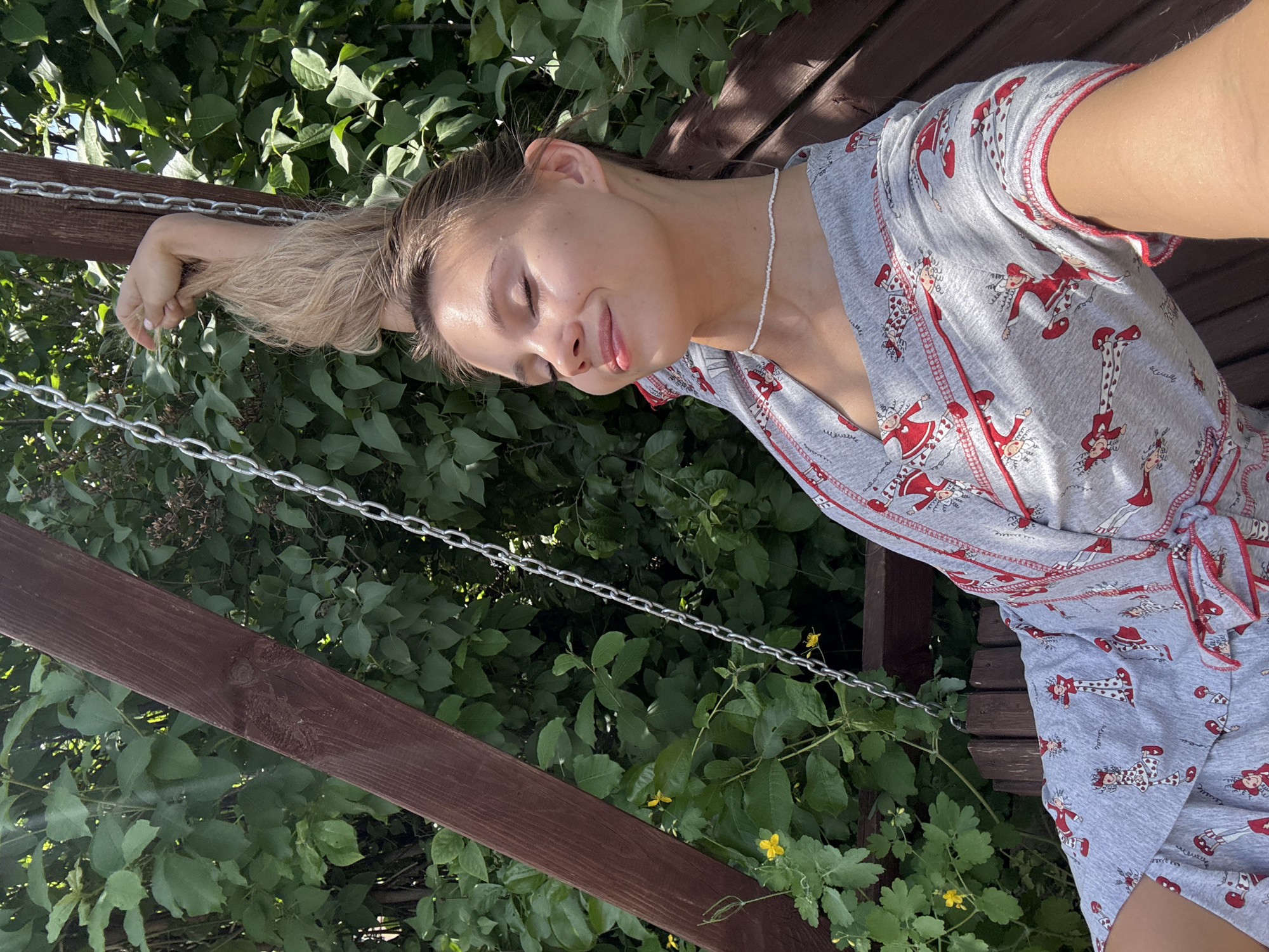 A young woman is resting on a wooden swing surrounded by green foliage, likely in a garden or park. She is wearing a grey patterned pajama set with red accents and a white beaded necklace. Her hair is blonde and pulled back in a ponytail. The swing is suspended by metal chains, and the background consists of dense greenery. The location is in the city of Chaikovsky, Russia.