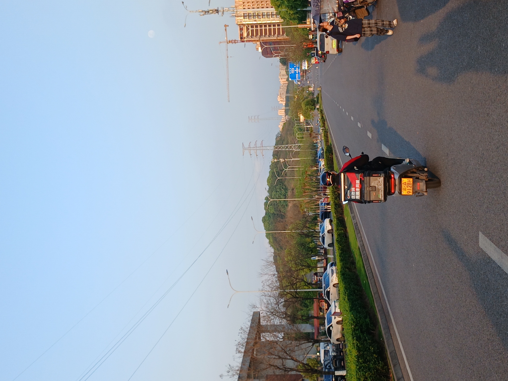 The image shows a street in the Shapingba District, China. A person on a scooter with a delivery box is riding on the right side of the road. Parallel parked cars line the left side of the road, separated by a median strip of greenery. Above the median are utility poles and green trees. In the distance are high-rise buildings and construction cranes. A person is lying on the sidewalk on the other side of the road, near some vehicles. The sky is light blue with some utility lines visible.