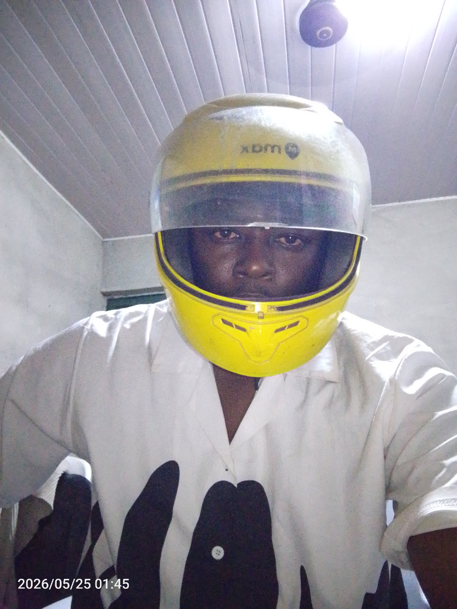 A close-up shot features a person wearing a yellow full-face motorcycle helmet with a clear visor. The helmet has a black stripe detail and the "XDM" logo visible on the top. The person's face is visible through the visor.
The person is wearing a white shirt with black splotches or patterns and visible buttons. The background consists of a tiled ceiling with a light fixture.
The timestamp "2026/05/25 01:45" is present at the bottom of the image.
No specific location within Lokoja, Nigeria, is discernible from the image.