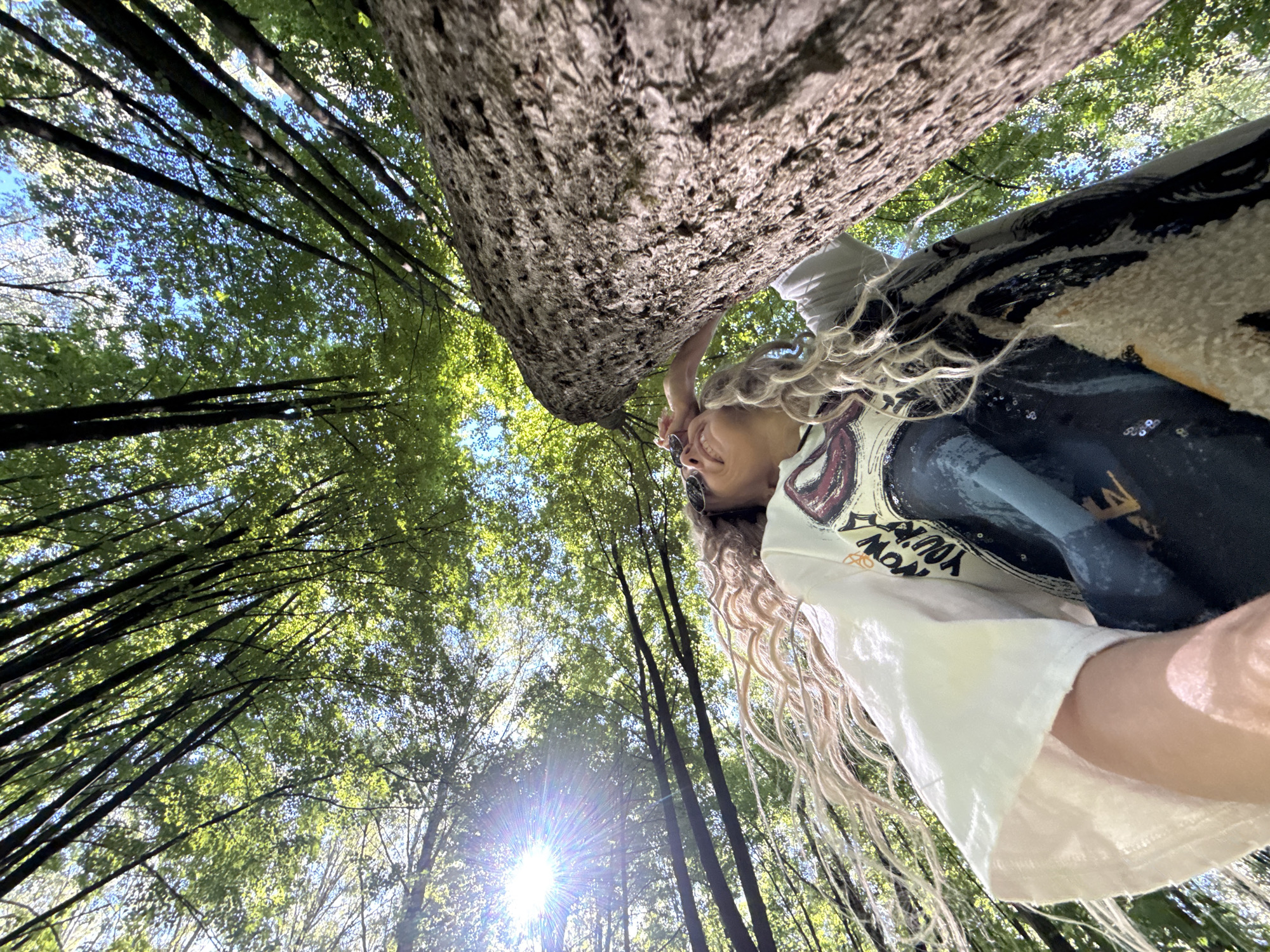 The image presents a forest scene in Rusanovo, Russia, captured from an extreme low-angle perspective looking directly upwards into a dense canopy of deciduous trees. The upper two-thirds of the frame are dominated by vibrant green leaves, interwoven with dark, slender tree branches, through which patches of clear blue sky are visible. A prominent sunburst lens flare is visible in the upper-left quadrant, indicating direct sunlight.
In the lower-right foreground, a large, vertical tree trunk with textured, rough gray-brown bark occupies a significant portion of the frame. In the lower-mid frame, an individual is partially visible, oriented in profile, looking upwards and slightly to the right. The individual has voluminous, wavy, light-colored hair that cascades down their left side. They are wearing dark, circular sunglasses, and their facial expression suggests a slight smile. The individual is dressed in a white short-sleeved t-shirt worn over a darker garment, which features a partially visible graphic design and text starting with "NOW YOU'RE". The individual's right hand is raised, with fingers lightly touching their temple near the sunglasses. The person appears to be positioned in close proximity to the large tree trunk on the right.