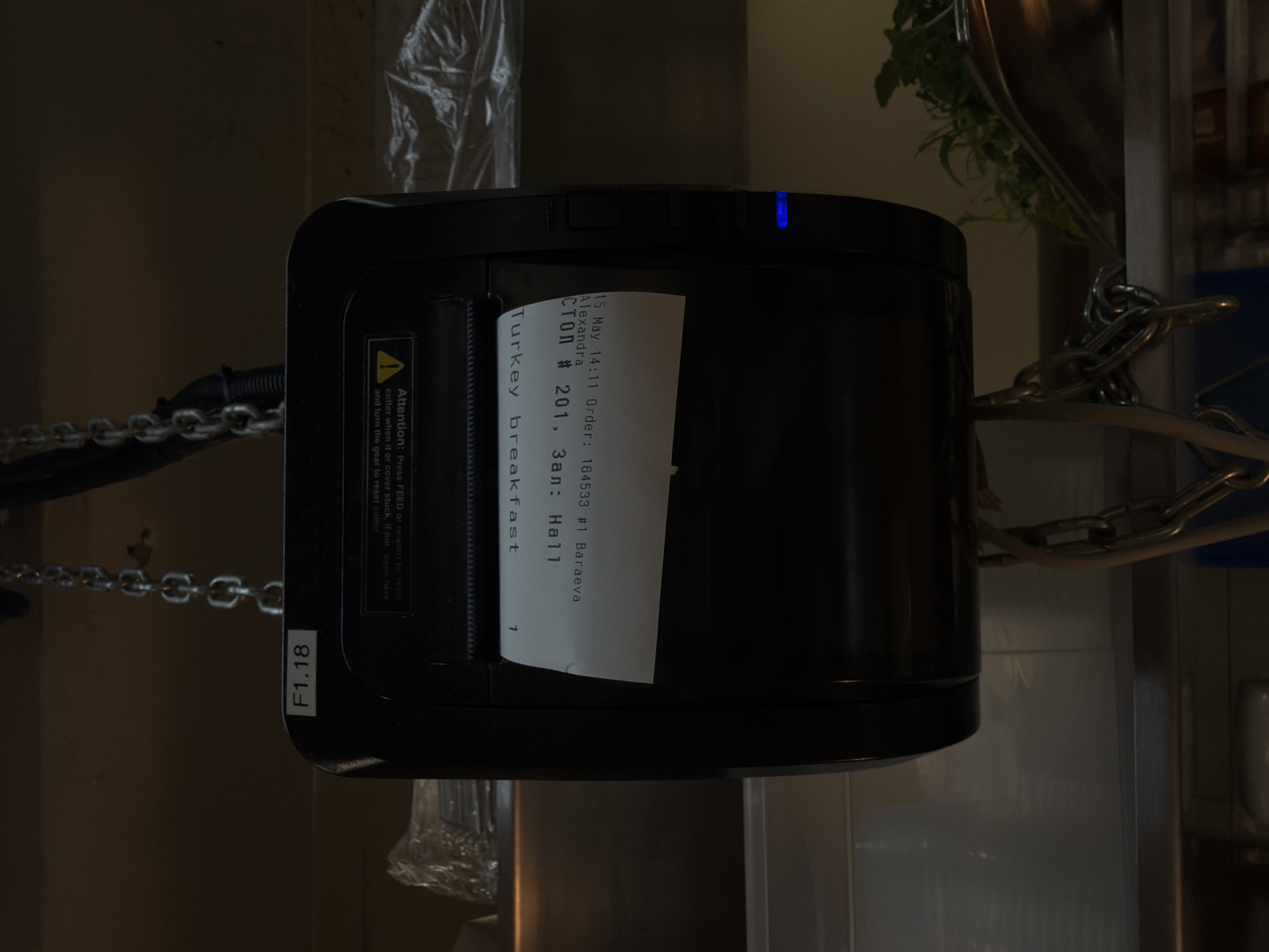 The image shows a thermal printer with a receipt partially printed. The receipt displays "15 May 14:11 Order: 164533 #1 Baraeva Alexandra СТОЛ # 201, Зал: Hall Turkey breakfast" along with a warning about the cutter mechanism. The printer is black with a blue light indicator. It's suspended by chains and located in what appears to be a kitchen environment with stainless steel surfaces, plastic wrapping, and a kitchen utensil holder visible in the background. The "Baraeva Alexandra" on the receipt suggests a location in Belgrade, Serbia, as Baraeva is a common surname in the region and "Alexandra" is a common first name. The text "СТОЛ # 201, Зал: Hall" indicates that the order is for table number 201 in Hall, likely within a restaurant or cafe.
