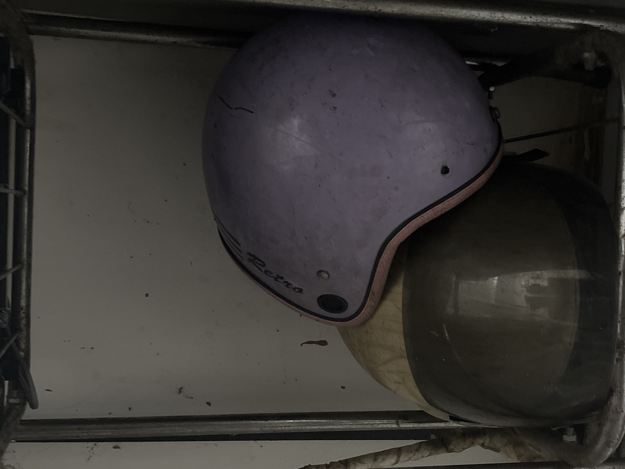 A high-angle, medium shot shows two helmets stacked on a shelf. The helmet on top is lavender with pink trim and a black circular emblem. Text on the helmet, partially visible, reads "Retro". The helmet below is dark brown with a reflective visor. The shelf appears to be made of metal and is dusty, as is the wall behind the helmets. The scene is dimly lit, suggesting an indoor storage area. The location of this image is a shelf in an unknown location in North Jakarta, Indonesia.
