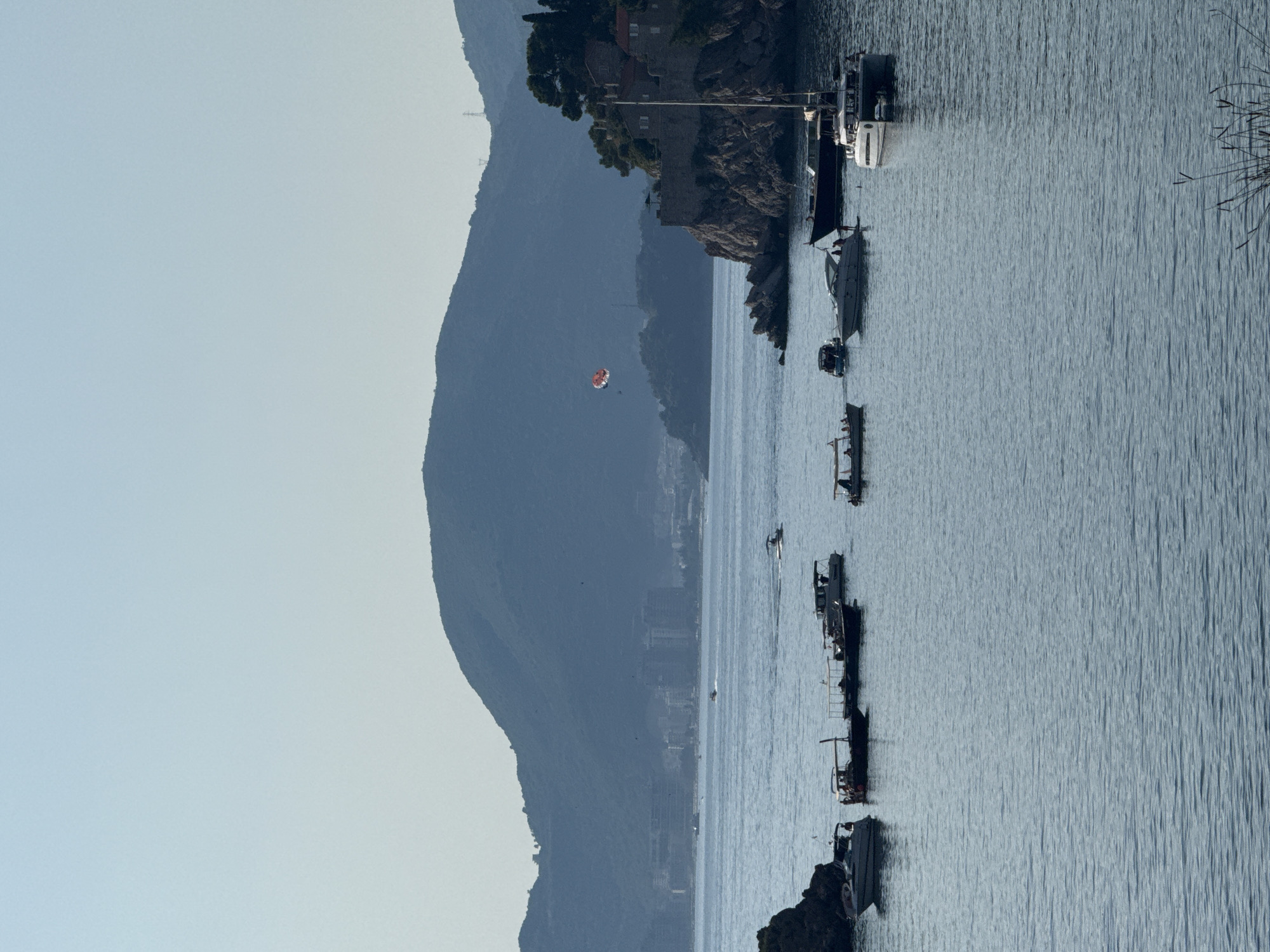 The image captures a serene coastal scene in Sveti Stefan, Montenegro, under a clear, pale blue sky. The foreground is dominated by the calm, blue sea, with gentle ripples reflecting the light. Several boats are scattered across the water. On the left, a white speed boat is moored. To its right, a longer, dark boat with a canopy is visible, followed by another boat of similar design. Further to the right, a sleek white speed boat is anchored, with two individuals on board. The largest vessel in the middle ground is a white catamaran with a dark hull.
In the mid-ground, a rocky outcrop juts into the sea on the right, topped with ancient stone structures and some trees, indicative of the historic island of Sveti Stefan. A sailboat with a white hull and mast is docked near this outcrop. In the distance, a mountain range forms a hazy backdrop, with a city skyline of buildings visible along the coast at the base of the mountains. A red and white parachute is visible in the sky above the island. The overall impression is one of a tranquil day at sea, with leisure activities taking place against a picturesque natural and historical landscape.
