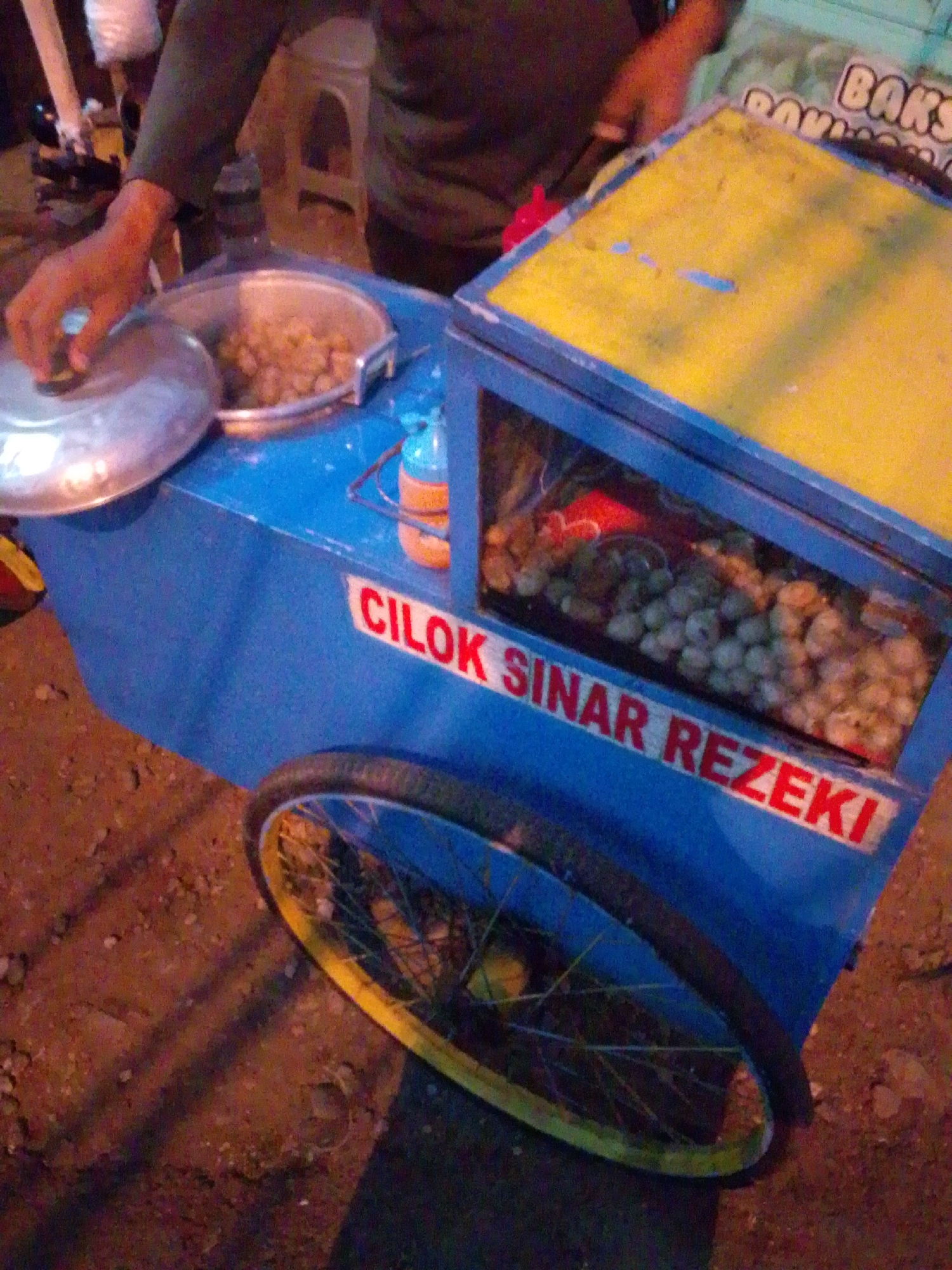 In Purworejo, Indonesia, a blue food cart with a yellow lid and the words "CILOK SINAR REZEKI" written in red is parked on a dirt surface at night. A person's hand is visible, reaching for a metal lid on a pot containing small round food items. Inside a glass display on the cart, more of these food items are visible, along with a red object. A bottle with orange liquid is also attached to the cart. A bicycle wheel with a yellow rim is attached to the cart's side. The background shows some unidentifiable structures and a sign that partially reads "BAKSO".