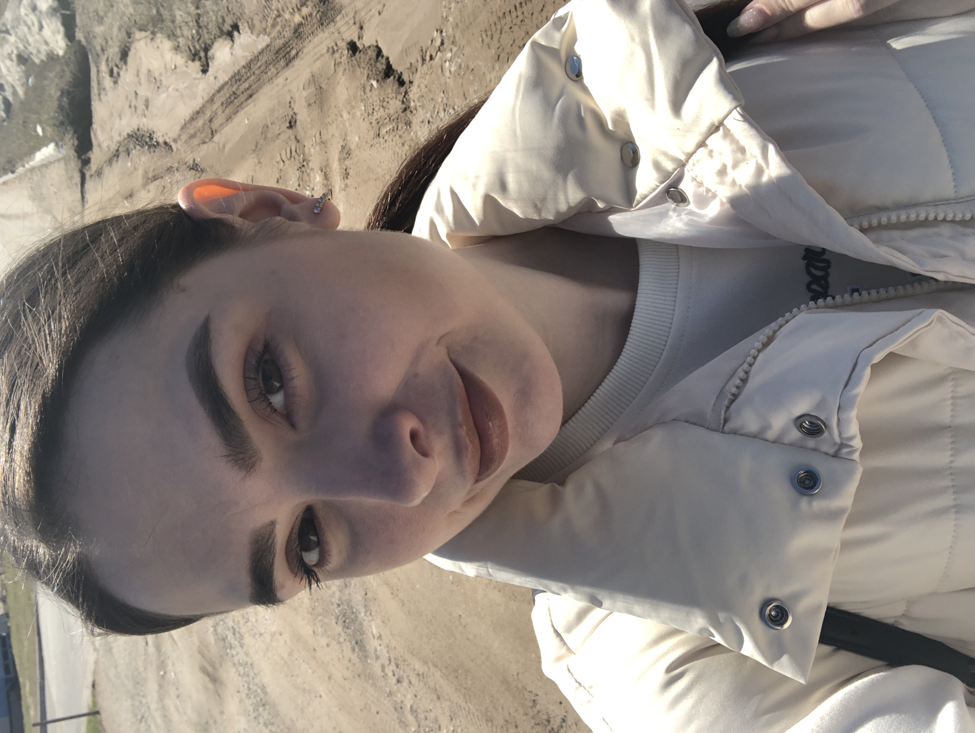A young woman is pictured in a close-up shot, likely taken outdoors. She has dark hair, arched eyebrows, and appears to be wearing light makeup, including lipstick. She is wearing a white quilted jacket over a white shirt. Her left ear is visible, showing a small earring.
The background appears to be a sandy or dirt area, possibly a beach or construction site. Tire tracks are visible in the dirt. It is possible the location is in the city of Noviy, Russia.