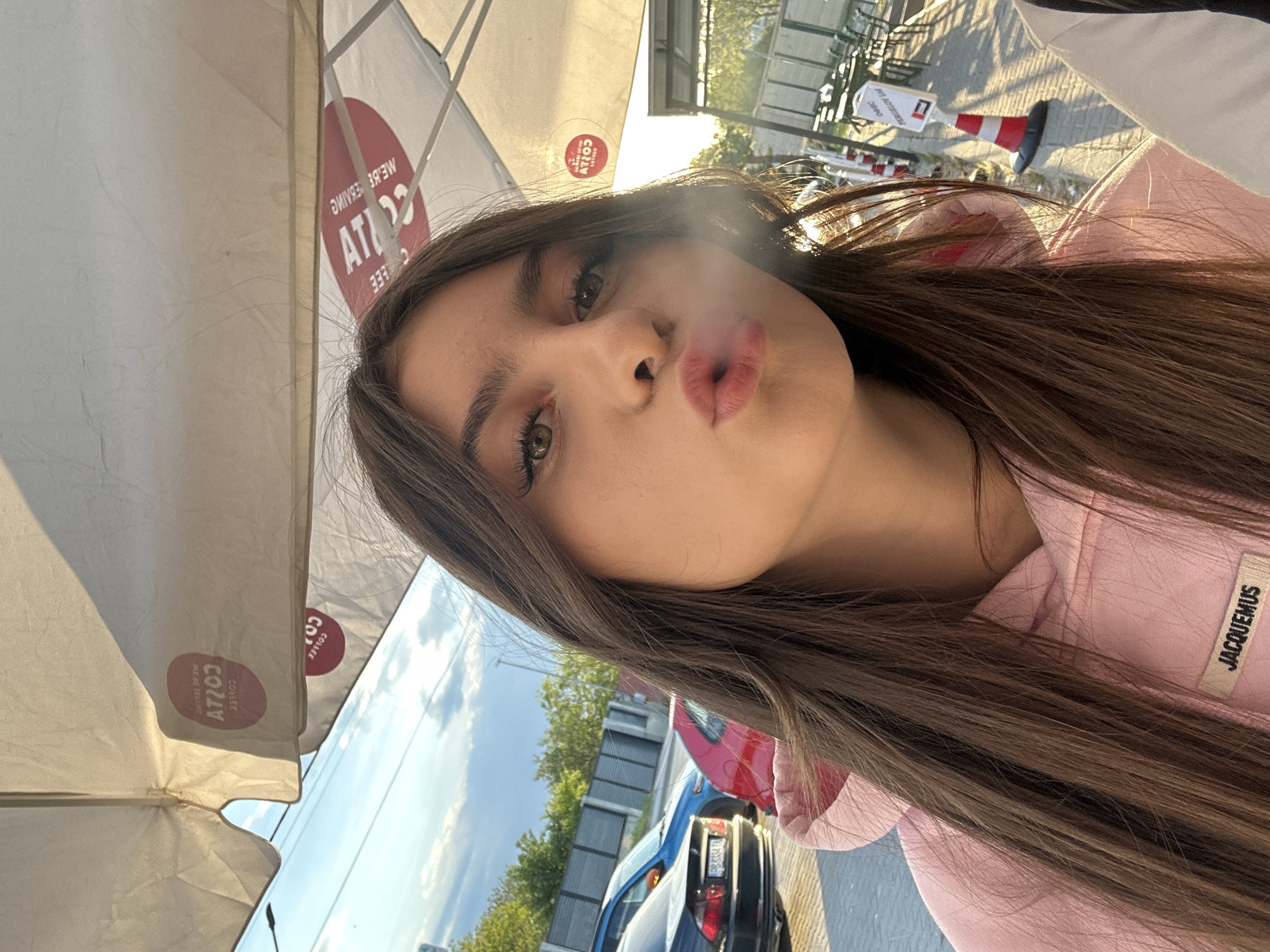 The image is a tilted selfie featuring a young woman with long, straight brown hair and light brown eyes, taken outdoors in Plovdiv, Bulgaria. Her lips are pursed as she exhales a misty plume, possibly vapor, looking directly at the camera with a posed expression. She wears a pink hooded sweatshirt, which has a white 'JACQUEMUS' label visible on the right side.
The scene depicts an outdoor cafe setting under bright daylight. Above and behind her, white patio umbrellas are partially visible, each featuring red circular logos with mirror-imaged text that strongly resembles 'COSTA COFFEE' branding. In the mid-ground, there are hints of outdoor seating and a red and white traffic cone. The background reveals a street or parking area with parked cars, including red and blue vehicles, and green trees under a clear, partially cloudy sky. The lighting suggests it is daytime, possibly late afternoon.