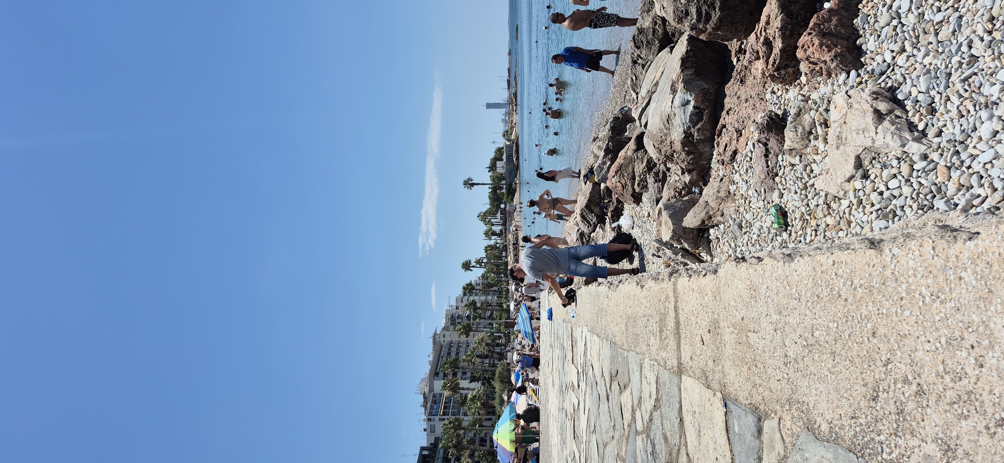 This is a full vertical shot of a beach scene in Paleo Faliro, Greece, on a clear, sunny day with a bright blue sky. The foreground features a concrete walkway bordering a rocky beach. A man in a grey t-shirt and blue denim shorts is standing on the walkway, facing away from the camera, holding a black bag. He appears to be interacting with something on the ground. Behind him, the rocky shore transitions to a sandy beach populated by numerous sunbathers and swimmers. Beach umbrellas in various colors dot the sandy area. In the background, several low-rise buildings, characteristic of a Mediterranean urban environment, are visible, interspersed with palm trees. Further out, the blue sea stretches to the horizon, where a tall skyscraper is faintly visible in the distance, along with more buildings and structures along the coastline. Several people are in the water, some wading and others swimming.