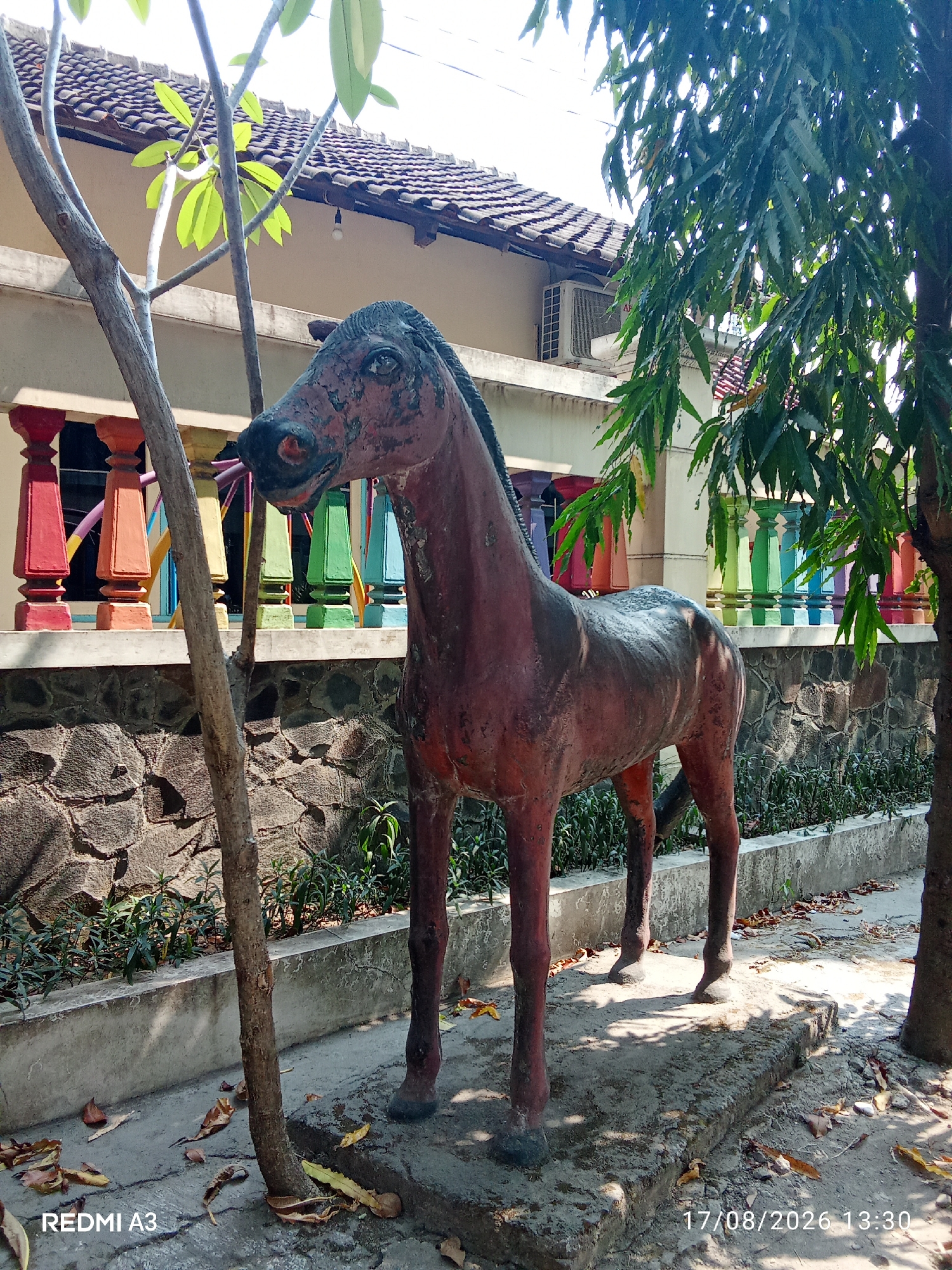 The image shows a statue of a horse in front of a building with a tiled roof and colorful railings. The horse statue is weathered and reddish-brown in color, standing on a concrete base. There are trees with green leaves on either side of the statue, casting shadows on the ground. The building has an air conditioning unit visible. The scene appears to be an outdoor area, possibly a garden or a yard, with fallen leaves scattered on the ground. The image also includes a watermark in the bottom left corner that reads "REDMI A3" and a date and time stamp in the bottom right corner that reads "17/08/2026 13:30". The presence of the REDMI A3 watermark suggests the photo was taken with a Xiaomi Redmi A3 smartphone. The building's architecture and the colorful railings might indicate a location in Indonesia, possibly within the city of Surakarta (Solo).