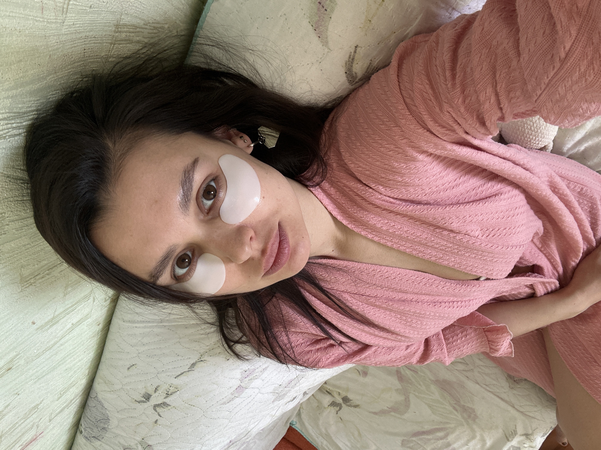A woman lies on a bed in a room that appears to be indoors. She has fair skin, brown eyes, and dark hair. Underneath her eyes, she wears white gel eye patches. She is dressed in a pink, woven, open-front robe. The bed is covered with a patterned white comforter. The location is presumably Chaikovsky, Russia. There is a white, knitted object near her hand.