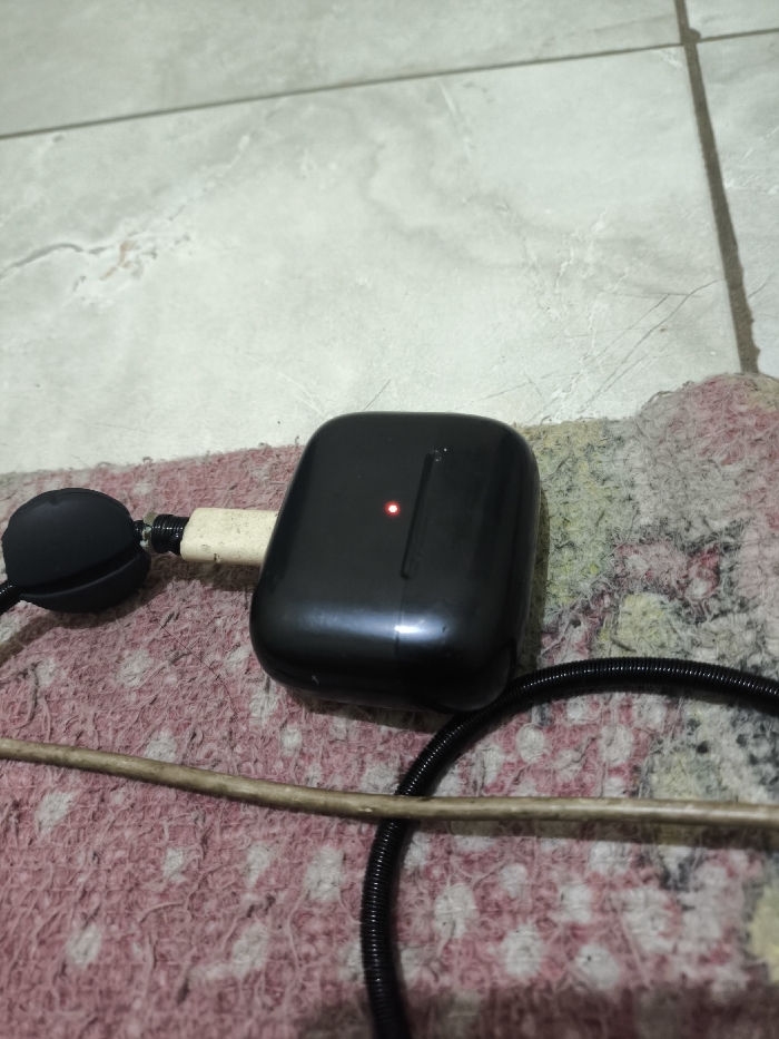 This indoor image, captured in Cibatu, Indonesia, provides a close-up view of a black charging case, likely for wireless earbuds. A small, illuminated red LED on its front surface indicates that the device is actively charging. To the left, a short, light-colored cable connects the case to a smaller, black spherical object, which could be an earbud or part of the charging setup.
These objects rest on a visibly worn, pinkish-red and off-white patterned fabric, resembling a mat or rug. A slender, light brown wooden stick or rod lies horizontally across the lower foreground, partially obscuring a coiled black corrugated cable. The background reveals a light-toned tiled floor with dark grout lines, characteristic of an interior setting. There are no individuals present, and no discernible text appears within the frame. The lighting suggests an interior environment.