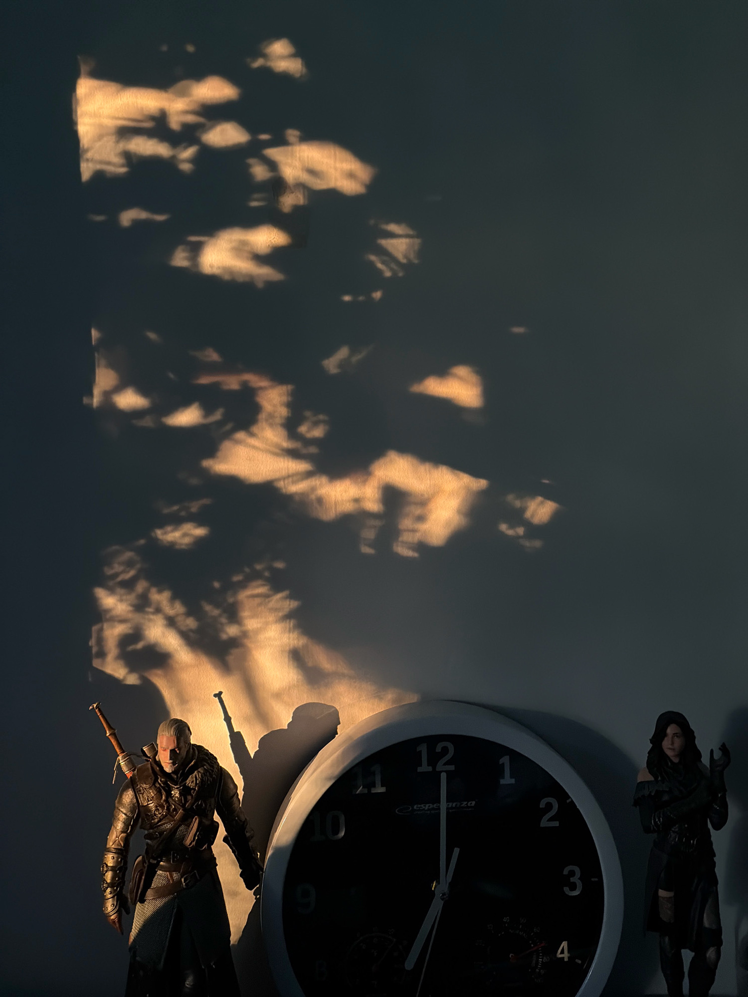 The image depicts a dimly lit interior scene with a wall as the primary background. Sunlight or artificial light casts dramatic shadows on the wall, creating an abstract pattern that resembles dappled foliage.
In the foreground, positioned on the left, is a male figurine dressed in medieval-style armor and carrying a sword on his back. The figurine appears to be a character from "The Witcher" series, possibly Geralt of Rivia. To the right of the figurine, and slightly behind it, is a large, dark-faced analog clock. The clock has white numerals and hands, and its face also displays smaller gauges and text, including the brand "esperanza".
Further to the right, standing behind the clock, is a female figurine, also dressed in dark, tactical-looking attire, reminiscent of a fantasy or game character. Both figurines are illuminated by a warm light source, likely originating from the left, which also creates distinct shadows of the figures and the clock on the wall.
There are no visible indications or elements within the image that suggest a specific location in the city of Oxentea, Moldova. The scene appears to be a domestic or display setting rather than an outdoor or public urban environment.