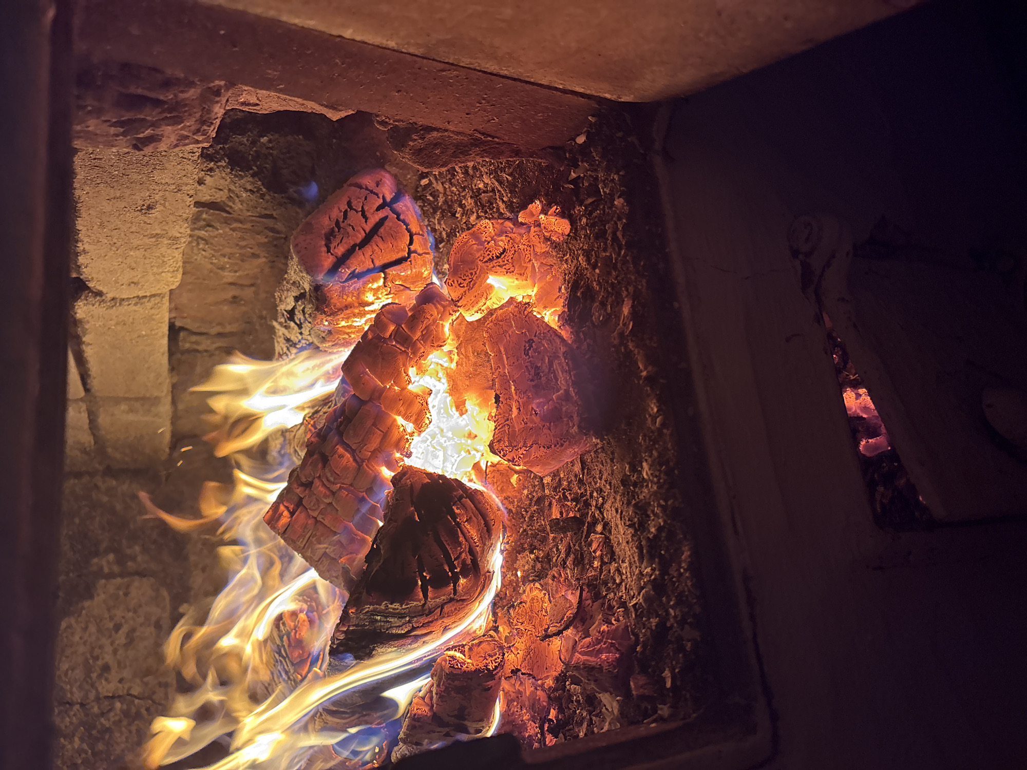The image shows a close-up view of a fireplace with burning logs and flames. The fire is bright orange and yellow, with hints of blue at the base of the flames. The logs are charred and cracked, with embers glowing beneath them. The fireplace is made of brick, and the surrounding area is dark and out of focus. There are no people or animals visible in the image. The scene is static and appears to be indoors. There is no information to suggest this scene is located in the city of Курган, Russia.