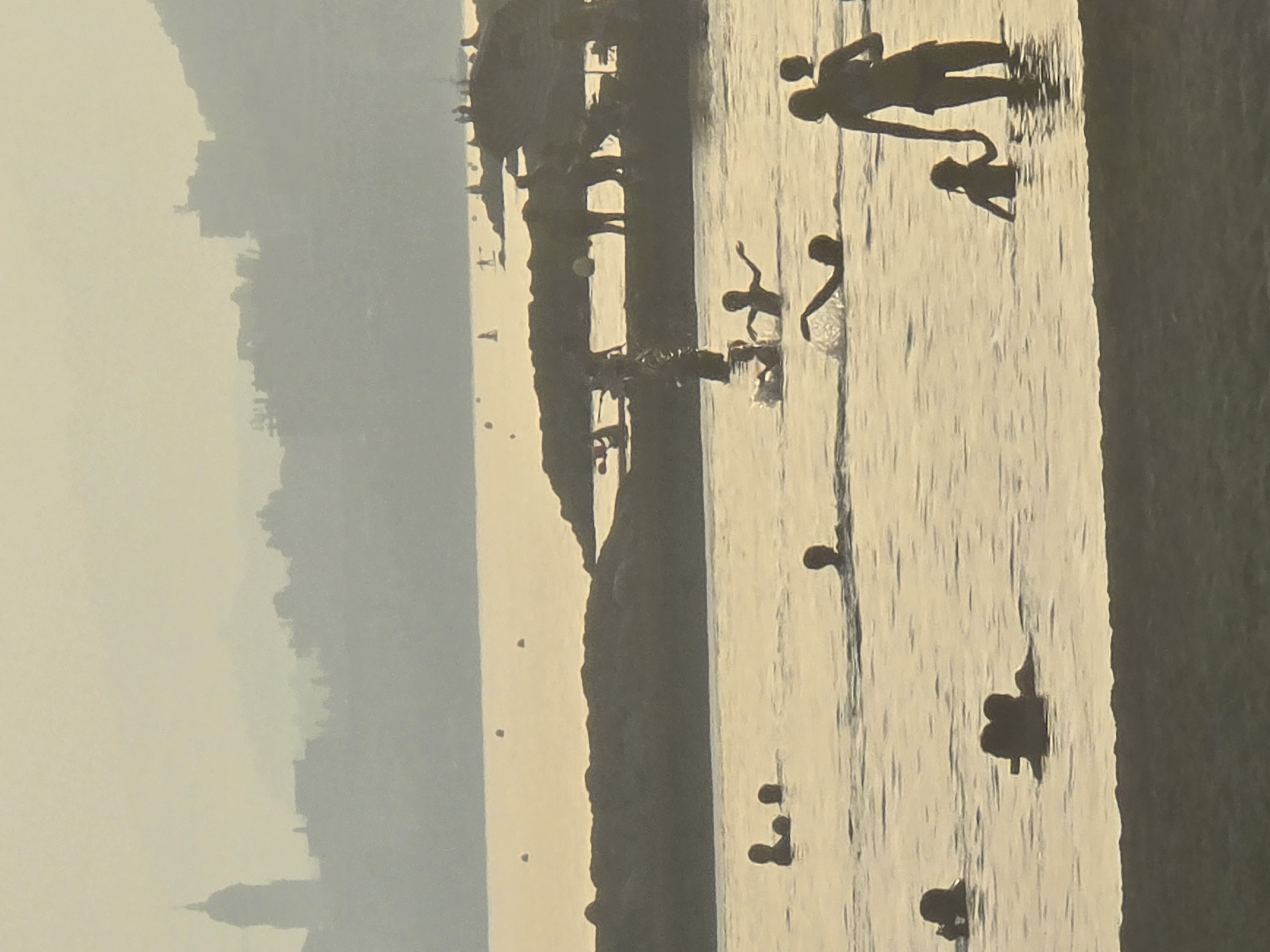 This is a hazy, silhouetted, medium shot of a beach scene in Málaga, Spain, likely taken at sunset or sunrise given the soft, golden light and diffused atmosphere. In the background, a city skyline with a prominent domed structure, possibly a cathedral or church tower, is visible, set against a hazy sky and distant hills. The middle ground features a rocky breakwater with several silhouetted figures standing or walking on it, and some appear to be on boats or paddleboards. The foreground is dominated by the calm sea, where numerous silhouetted figures, including adults and children, are swimming, playing, and wading. Some figures are interacting, such as a woman holding a child's hand, and children splashing. The water is calm with gentle ripples reflecting the muted sky. The overall impression is one of a peaceful, idyllic beach day.