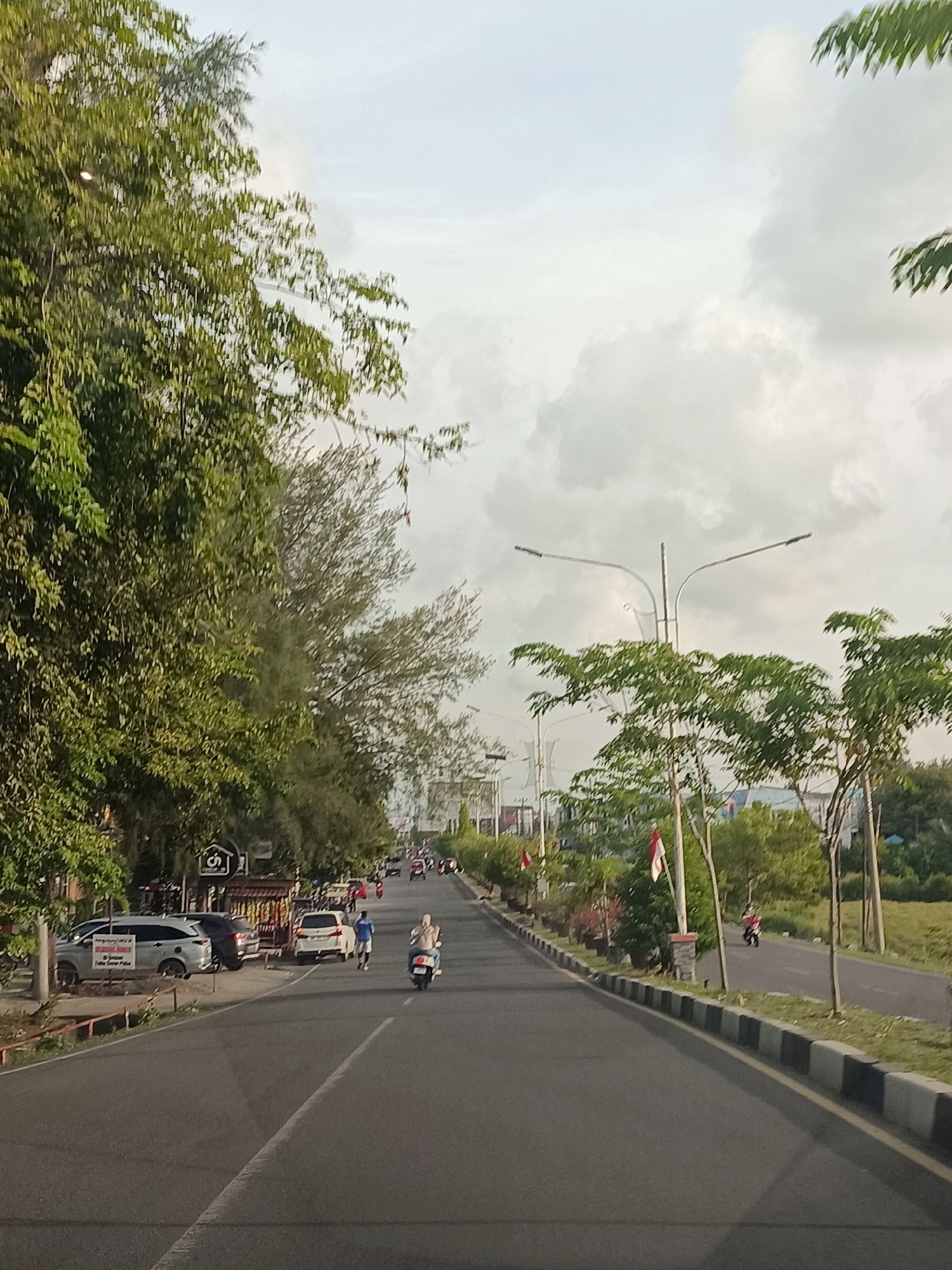 The image depicts a street scene in Sigli, Indonesia, under a cloudy sky. The road is paved and has white lane markings. On the left side of the road, there are several parked cars, including a white SUV and a darker vehicle. A few people are visible on the sidewalk and near the parked cars. Further down the road, a person is riding a scooter, and another person is walking.
The right side of the road features a sidewalk with some greenery and trees. There are streetlights along the road. In the distance, more trees and buildings are visible. Two small Indonesian flags are planted in the ground on the right side of the road. The overall atmosphere suggests a moderately busy street during the daytime.