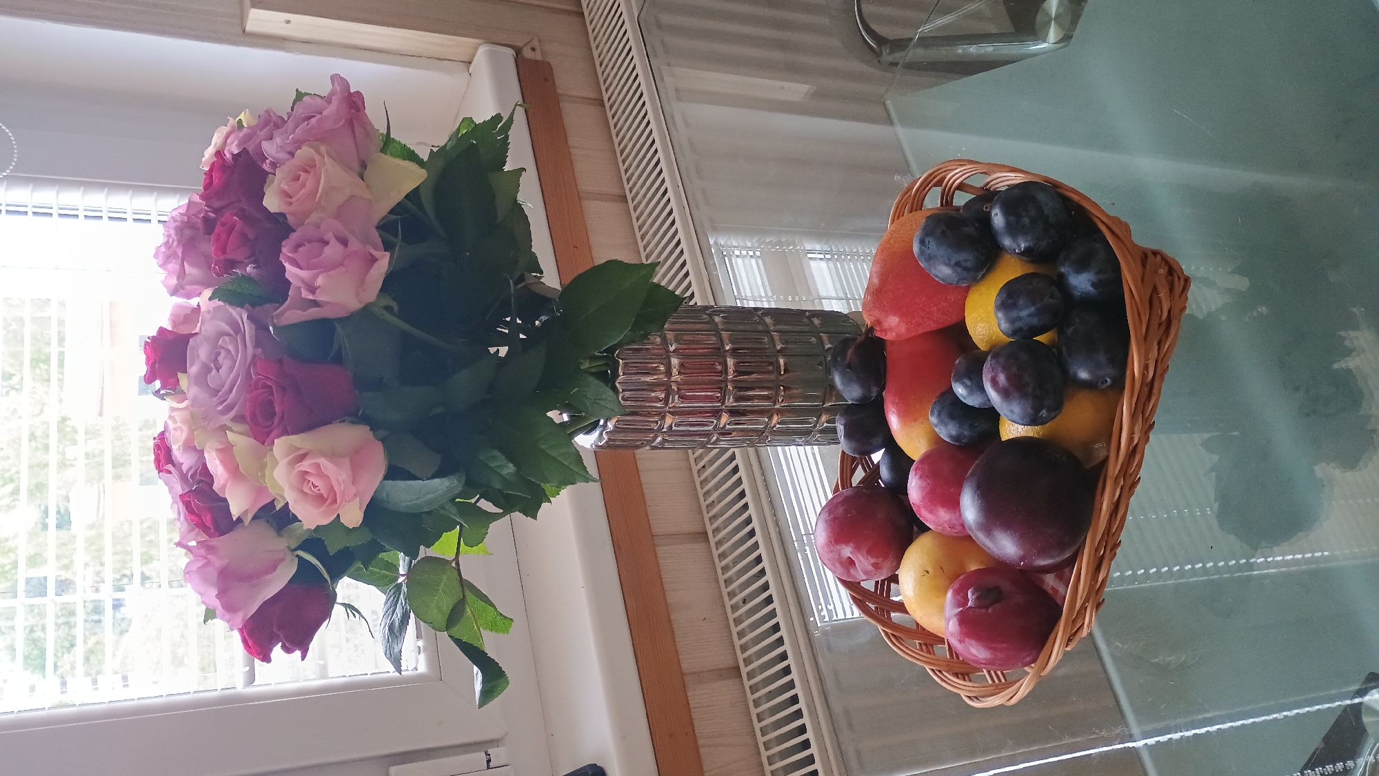 The image depicts a still life arrangement on a glass table, likely in a home in Sokolov, Czechia. In the background, a bouquet of roses in various shades of pink and red is displayed in a textured silver vase. The vase sits on a windowsill with blinds covering the window, suggesting an indoor setting. In the foreground, a woven basket is filled with a colorful assortment of fruits. The basket contains several dark purple plums, two yellow lemons, and what appear to be red plums or nectarines. The glass table reflects the objects above it, adding a layer of visual depth to the scene. The overall atmosphere is one of domestic tranquility.
