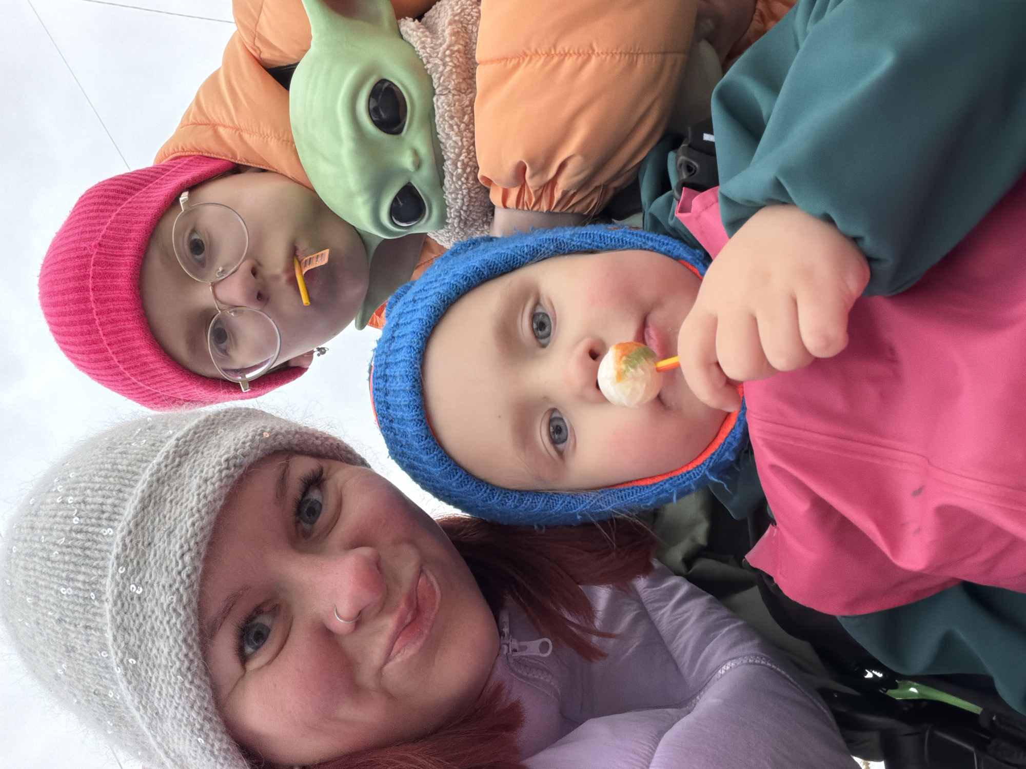 A low-angle selfie shows a woman wearing a light gray knitted hat, light purple puffy jacket, and a nose ring, next to a child wearing a blue beanie and green jacket, who is eating a lollipop. A girl wearing round glasses and a pink beanie with a yellow object protruding from her mouth is next to a Grogu mask with big black eyes and no mouth. All subjects are oriented towards the camera. The exact location in Chaikovsky, Russia, is not discernible from the image.